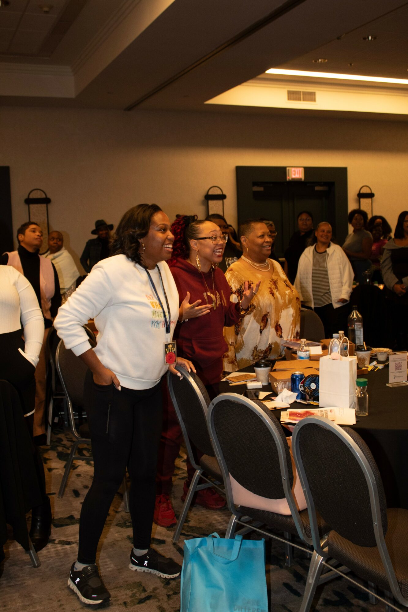 Group of people standing in a conference room, some with hands on hips, facing forward, with tables and chairs nearby.