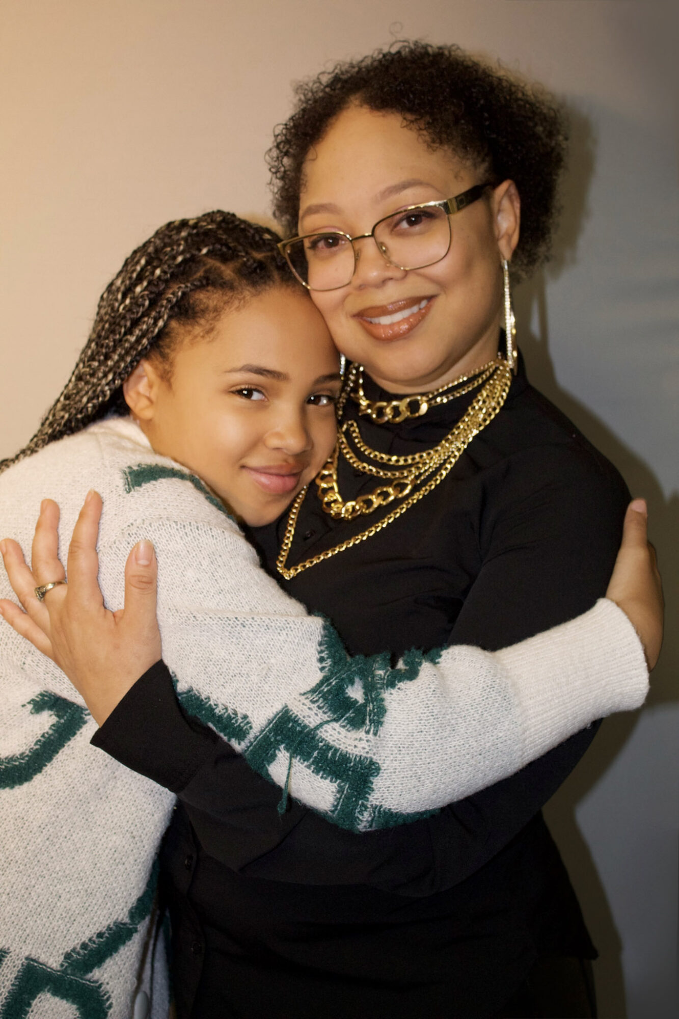Two people hugging, one with glasses and jewelry, the other with braided hair, smiling warmly.