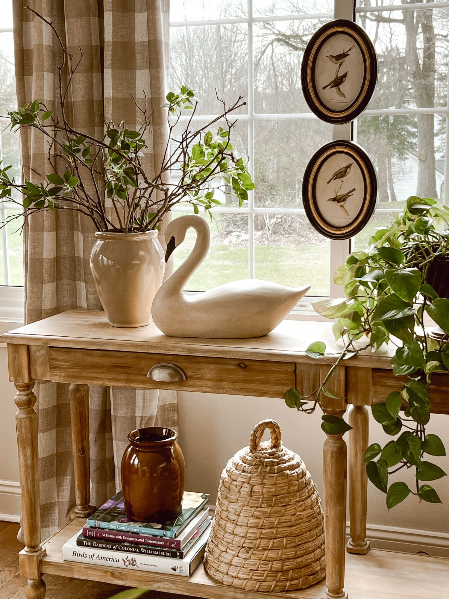 Wooden table with a vase of branches, a swan sculpture, and a plant near a window with curtains.