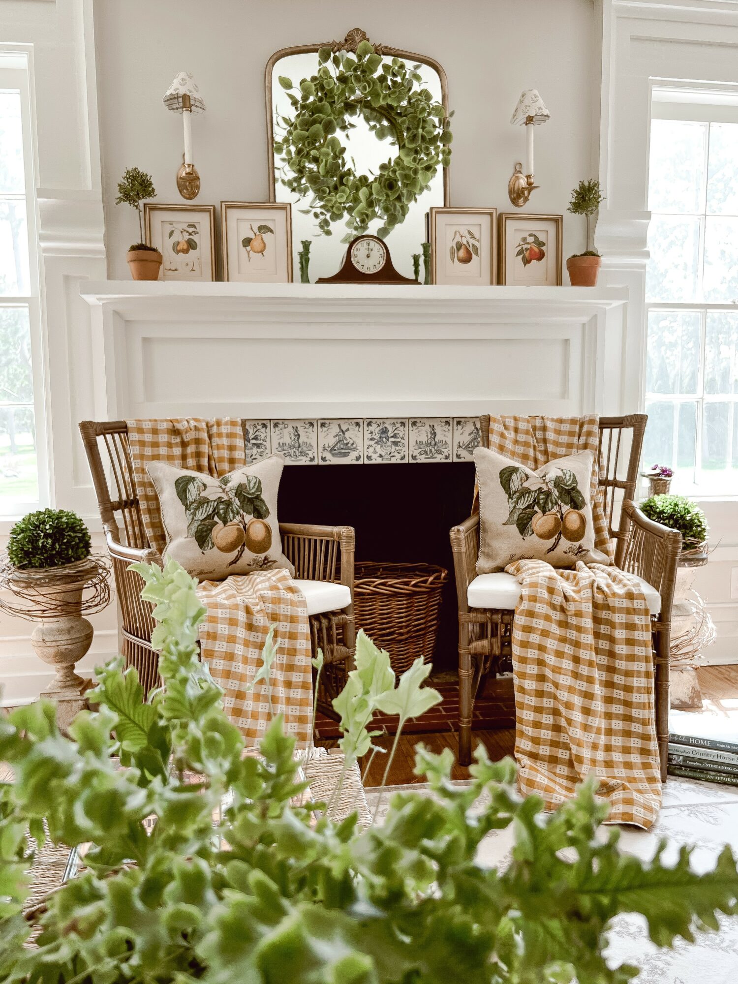 Living room with a white fireplace, decorated with a wreath, framed art, and potted plants, flanked by two windows.