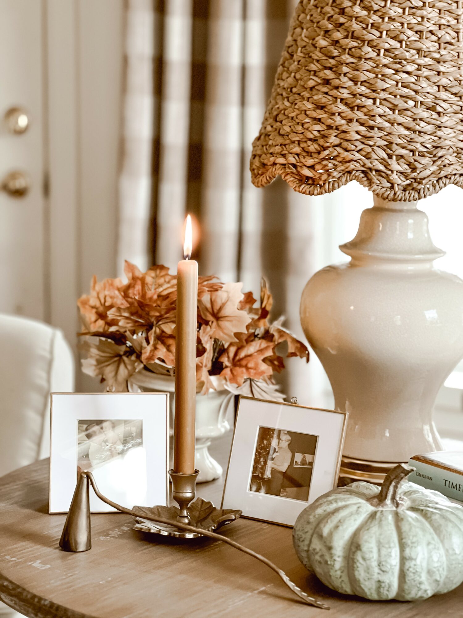 Decorative table with framed photos, a candle, a pumpkin, a vase with flowers, and a lamp with a woven shade.