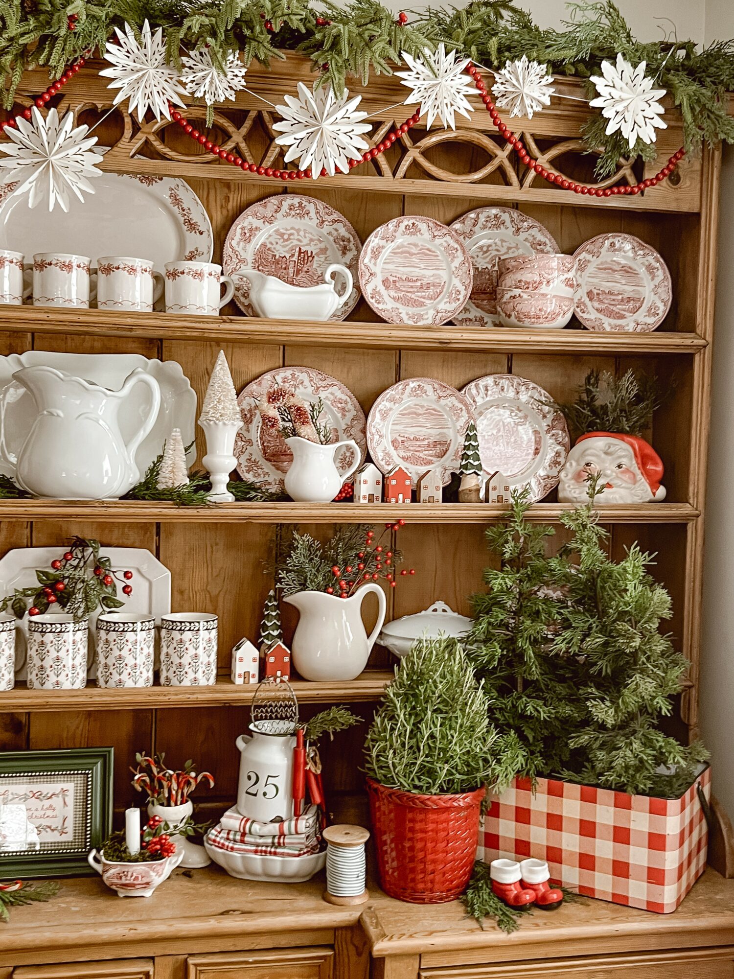 Wooden shelf with Christmas decorations, plates, teapots, and small trees, decorated with snowflakes and garlands.