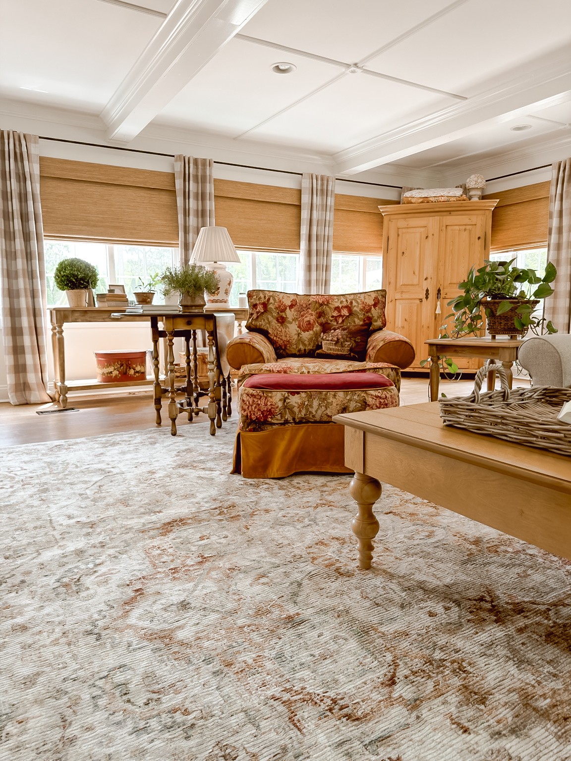Living room with a floral sofa, wooden furniture, large windows with curtains, and a carpeted floor.