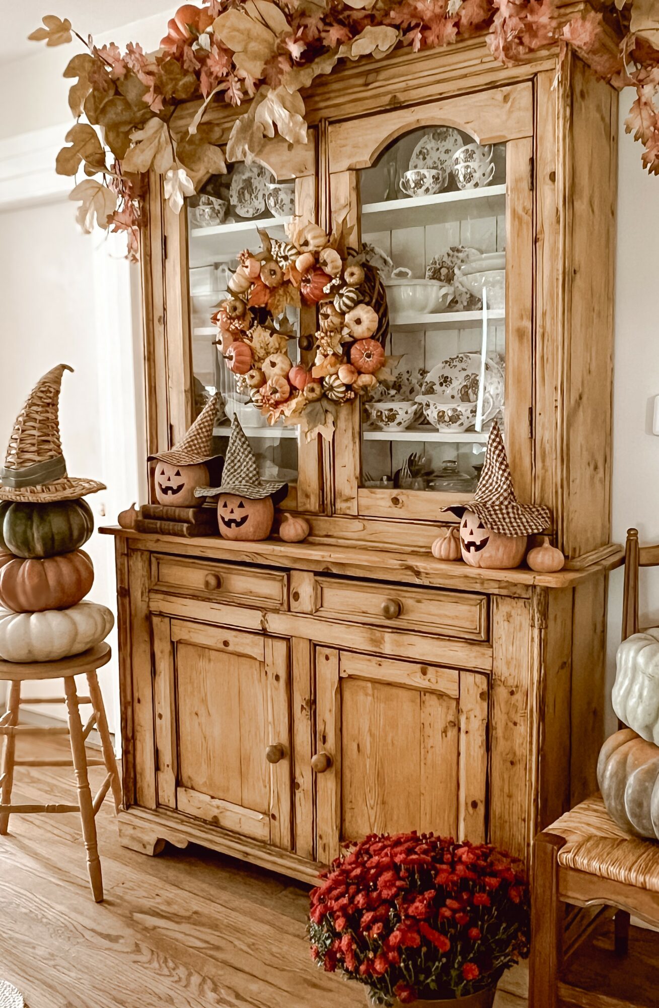 Wooden cabinet decorated with autumn-themed wreath, pumpkins, and floral arrangements, with stacked pumpkins and flowers nearby.
