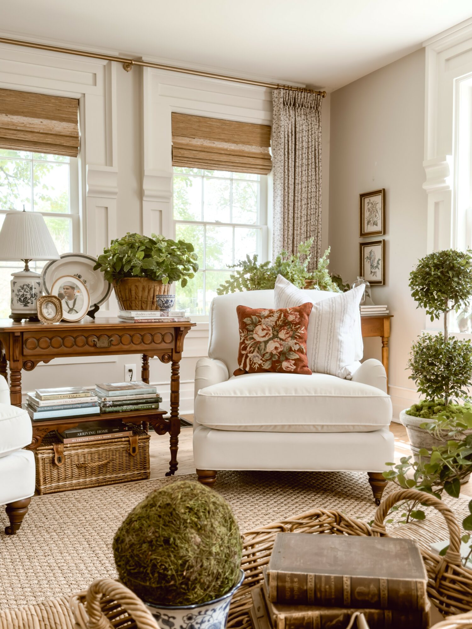Cozy living room with white armchair, wooden side table, plants, and large windows with curtains and blinds.