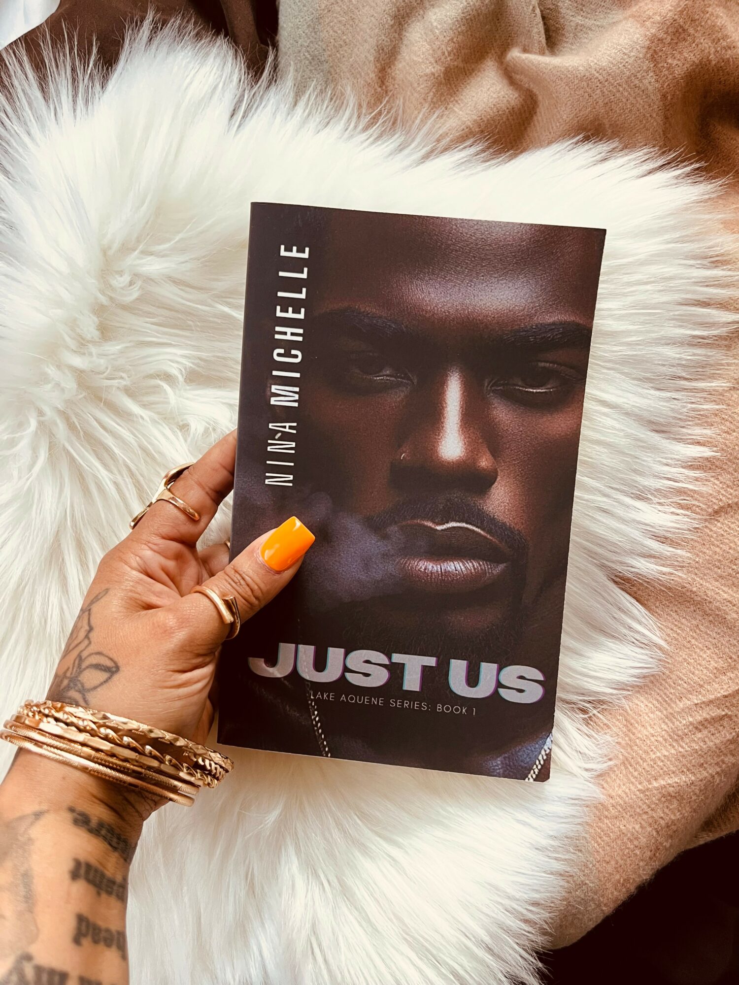 Hand holding a book titled 'Just Us' with a dark-skinned person's face on the cover, resting on a white furry surface.