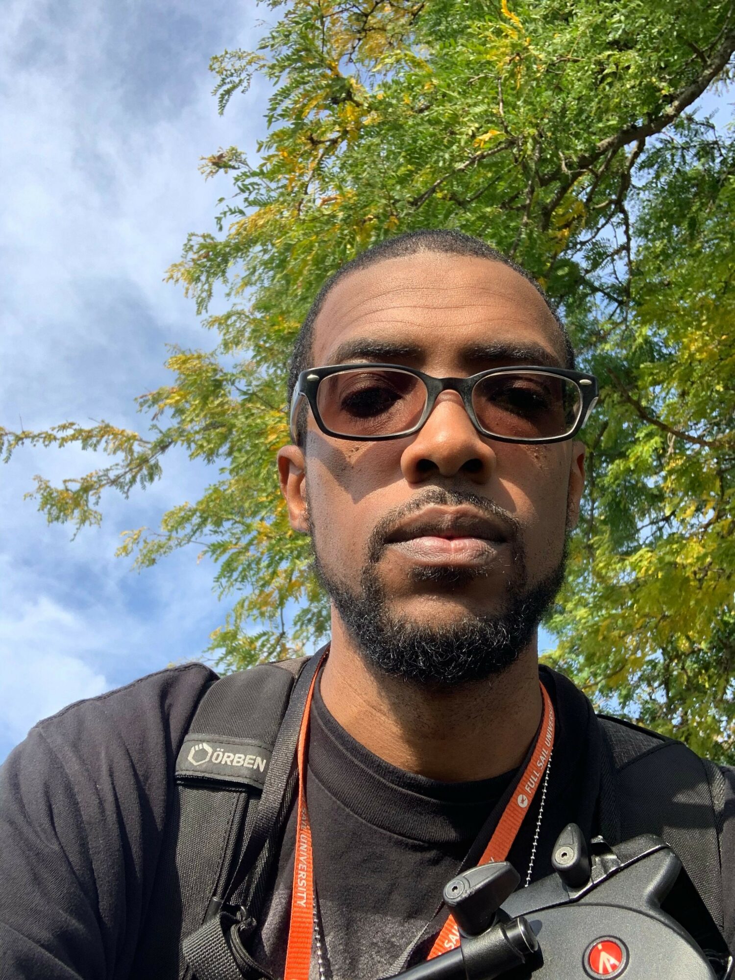 Man with glasses and a beard outdoors, trees and blue sky in background.