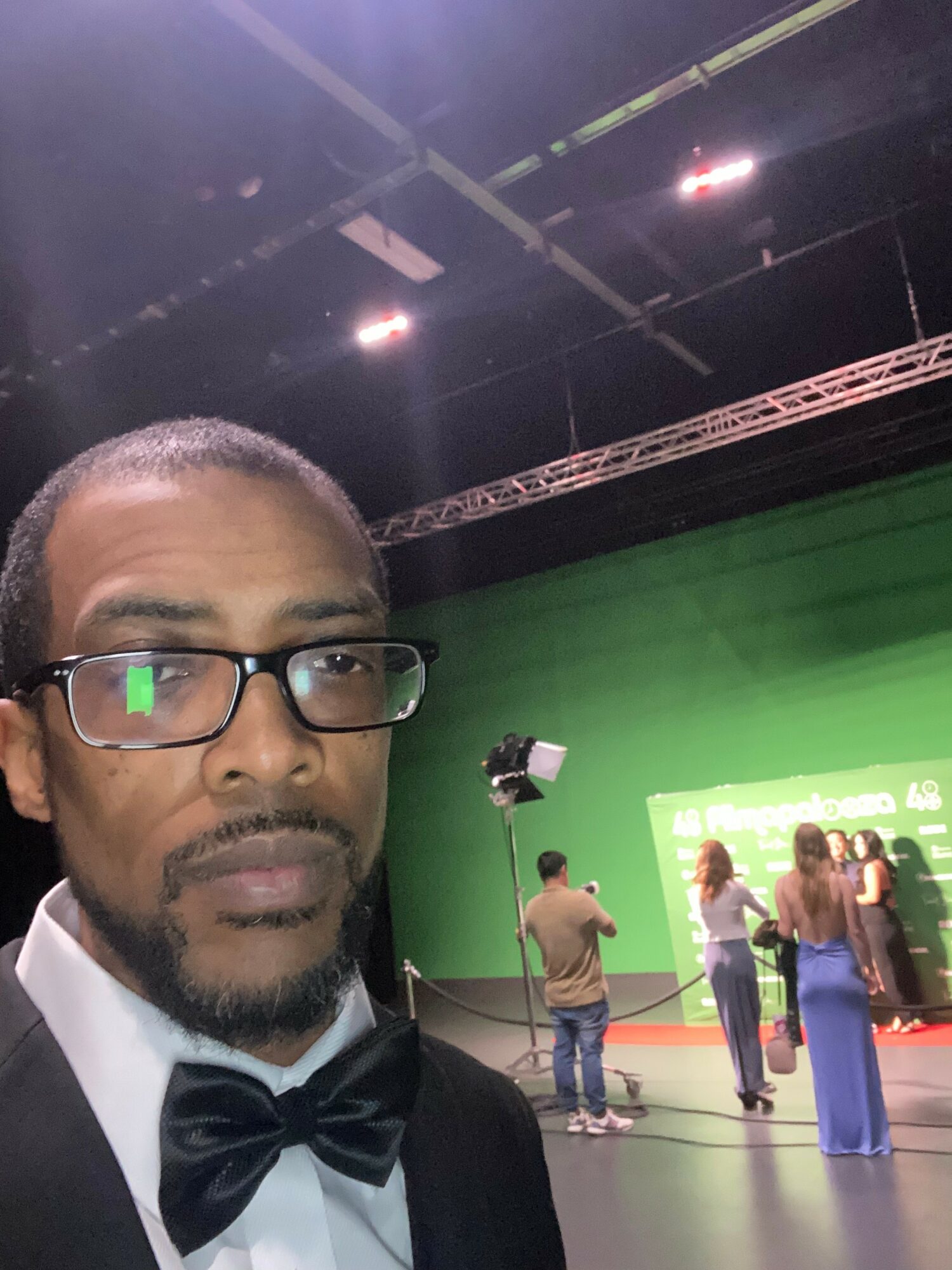 Man with glasses and bow tie taking selfie in studio with green screen and people in background