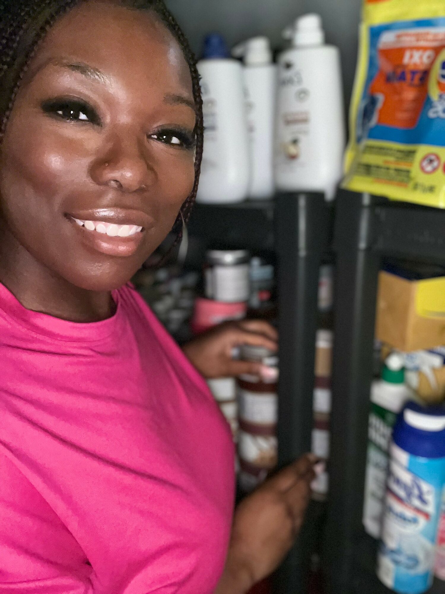 Woman in pink shirt smiling, standing near shelves with bottles and containers, holding a small item in her hand.