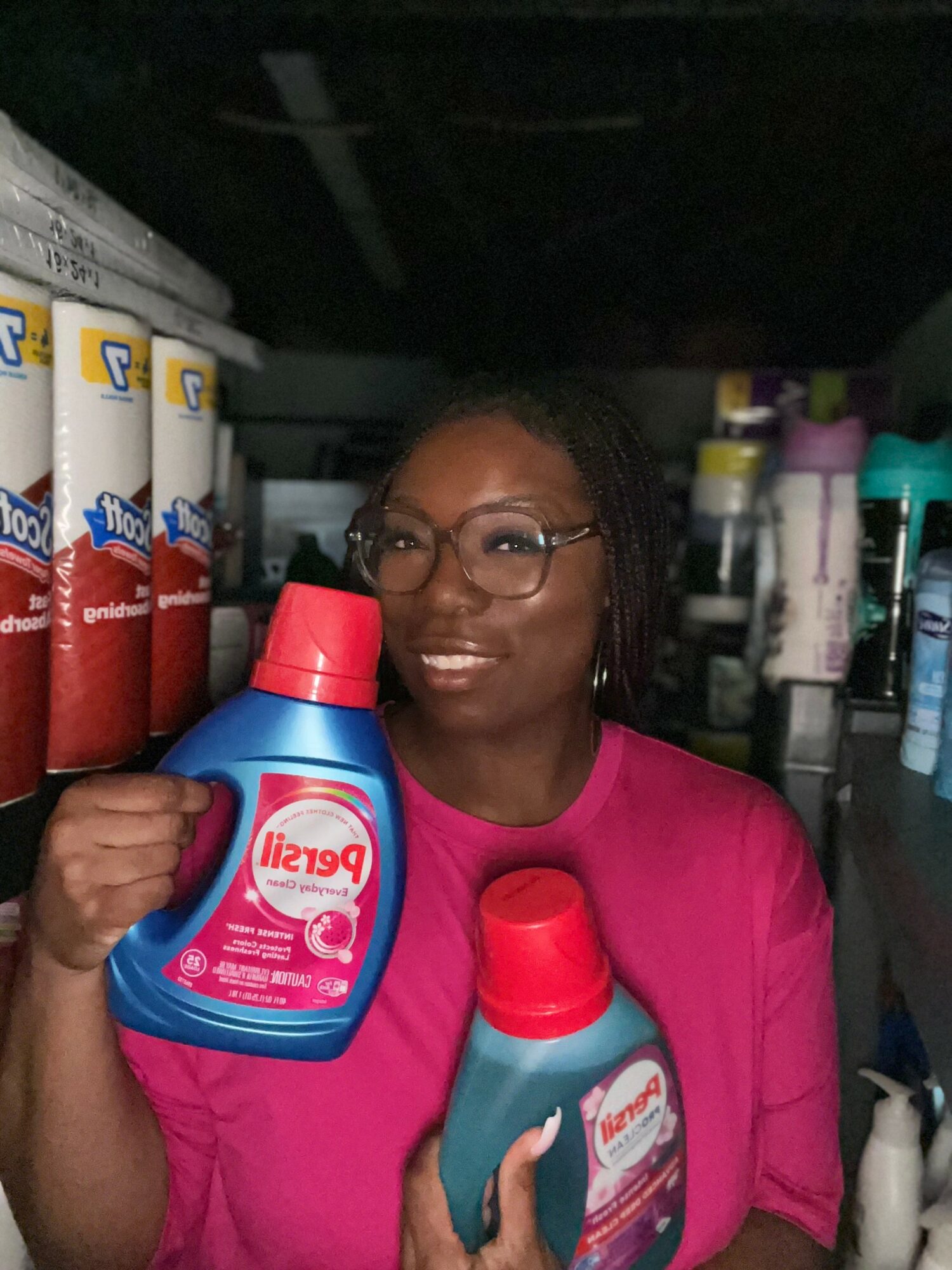 Woman with glasses holding two bottles of cleaning products, shelves with various bottles behind her.