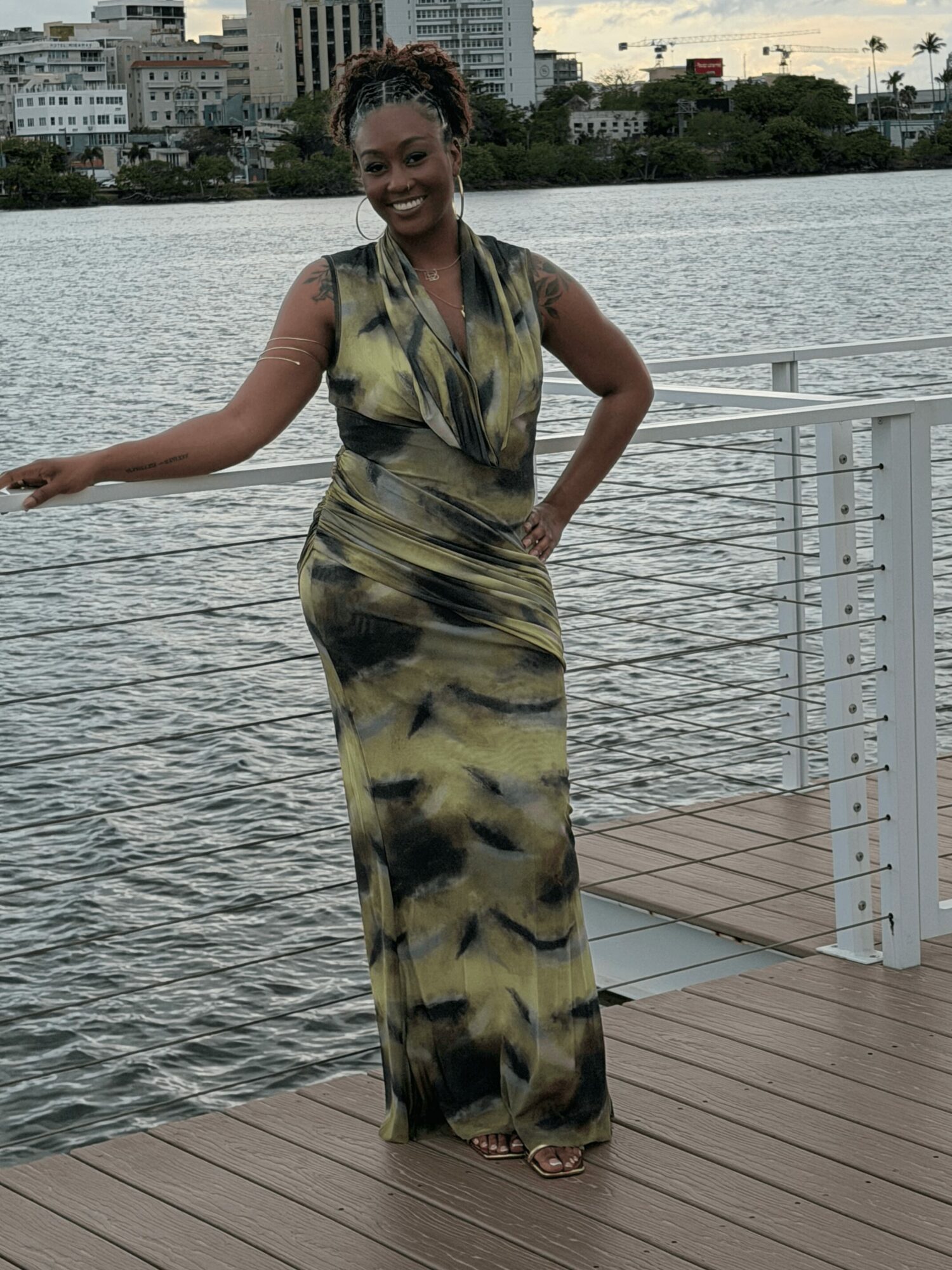 Woman in a long dress standing on a dock by water, smiling, with city buildings in background.