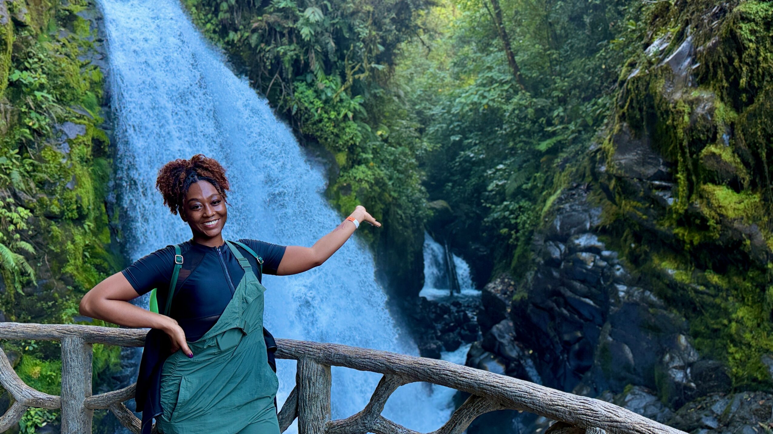 A woman with curly hair stands on a wooden railing in front of a waterfall surrounded by lush greenery.