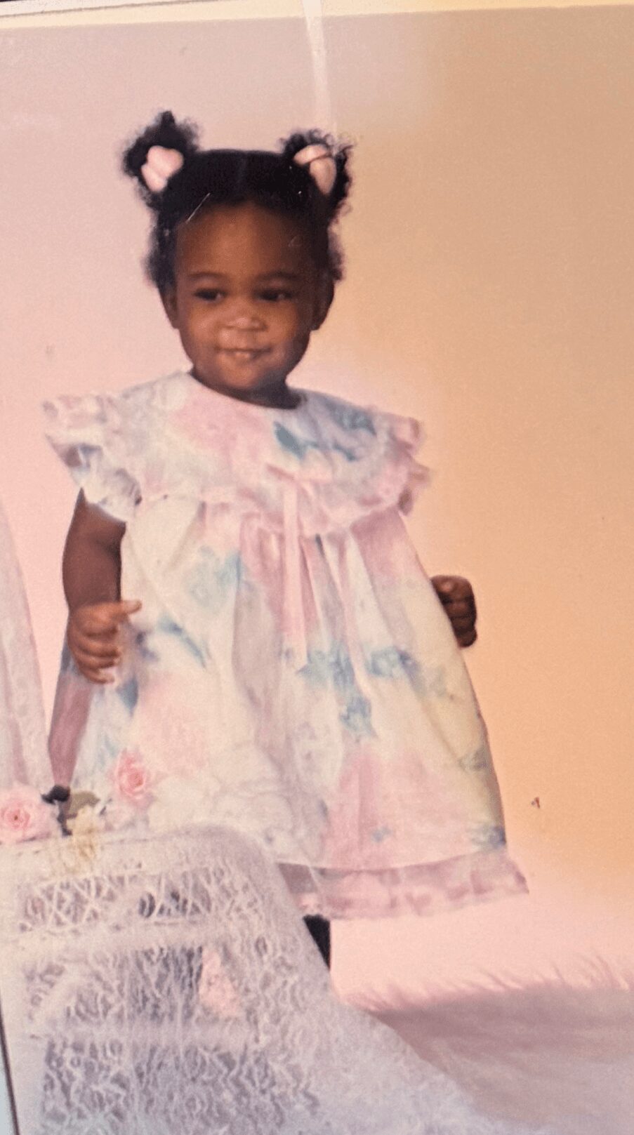 Young girl with curly hair styled in two buns, wearing a pastel-colored dress with ruffles, standing indoors.