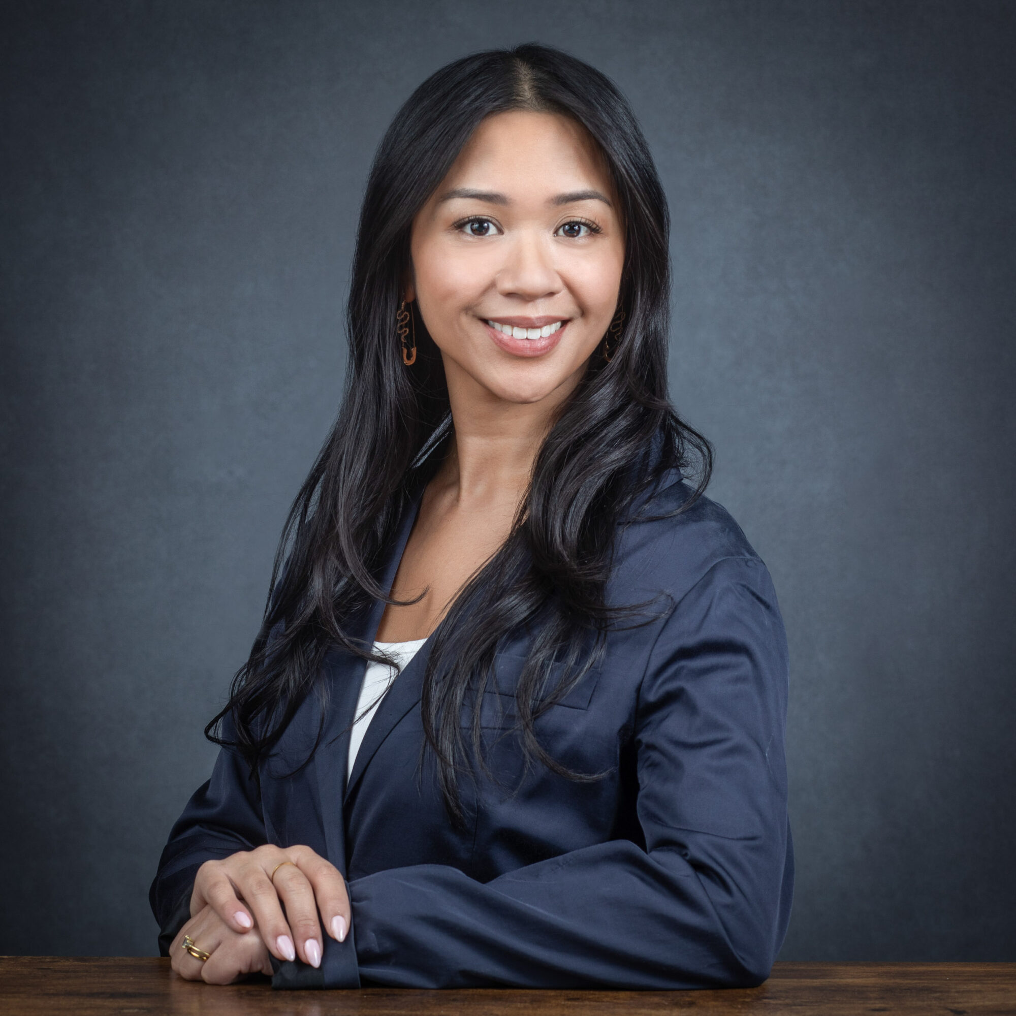 Young woman with long black hair smiling, wearing a navy blazer, sitting at a wooden table against a dark background.