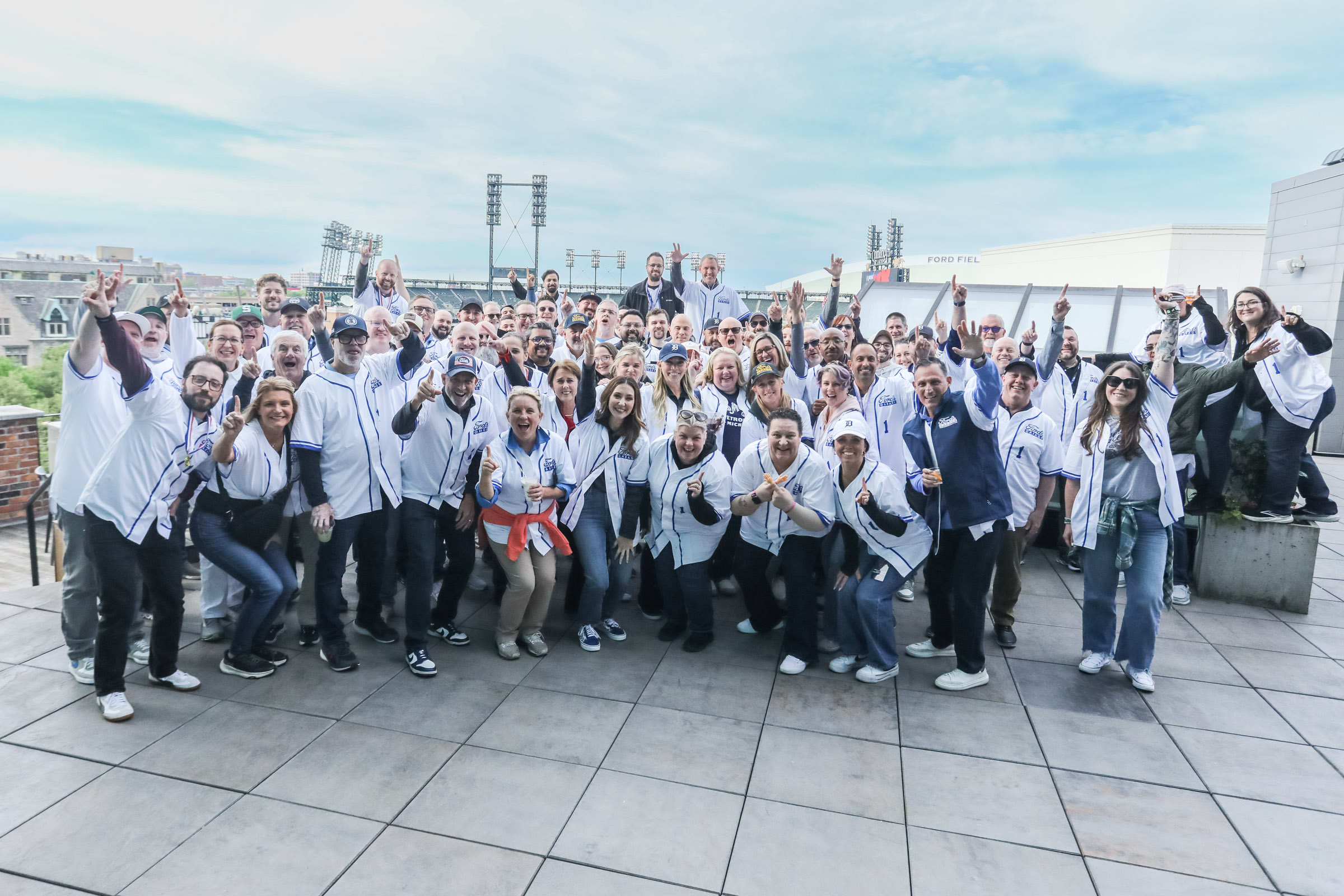Group of people on a rooftop, smiling and raising their hands, with cityscape and blue sky in background.