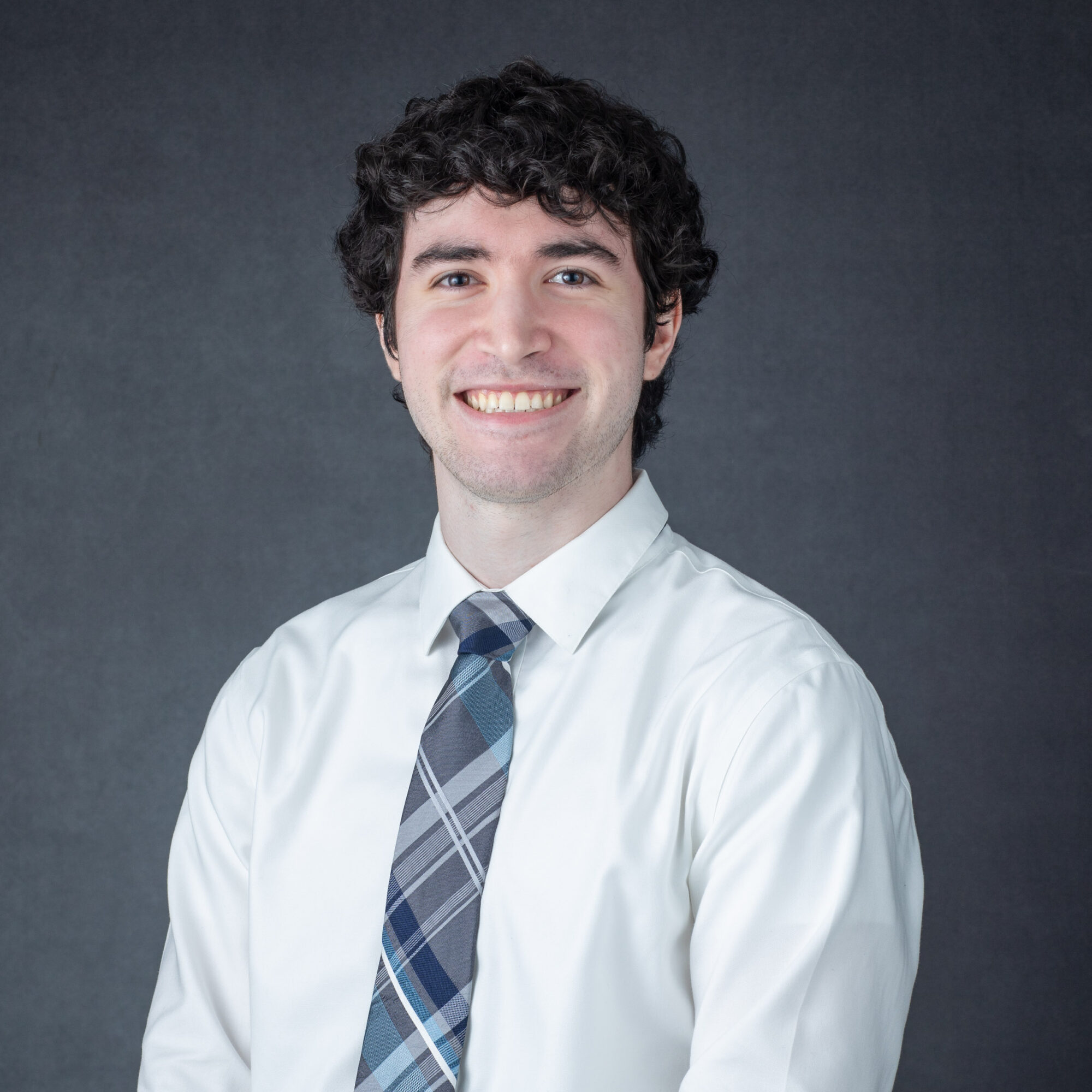 Young man with curly hair smiling, wearing a white shirt and a plaid tie, against a dark background.