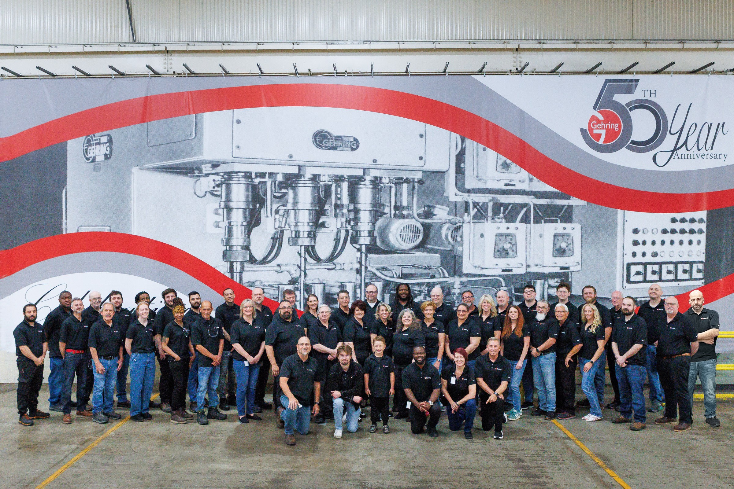 Group of people standing in front of a large industrial mural with red and gray design elements, celebrating a 50-year anniversary.