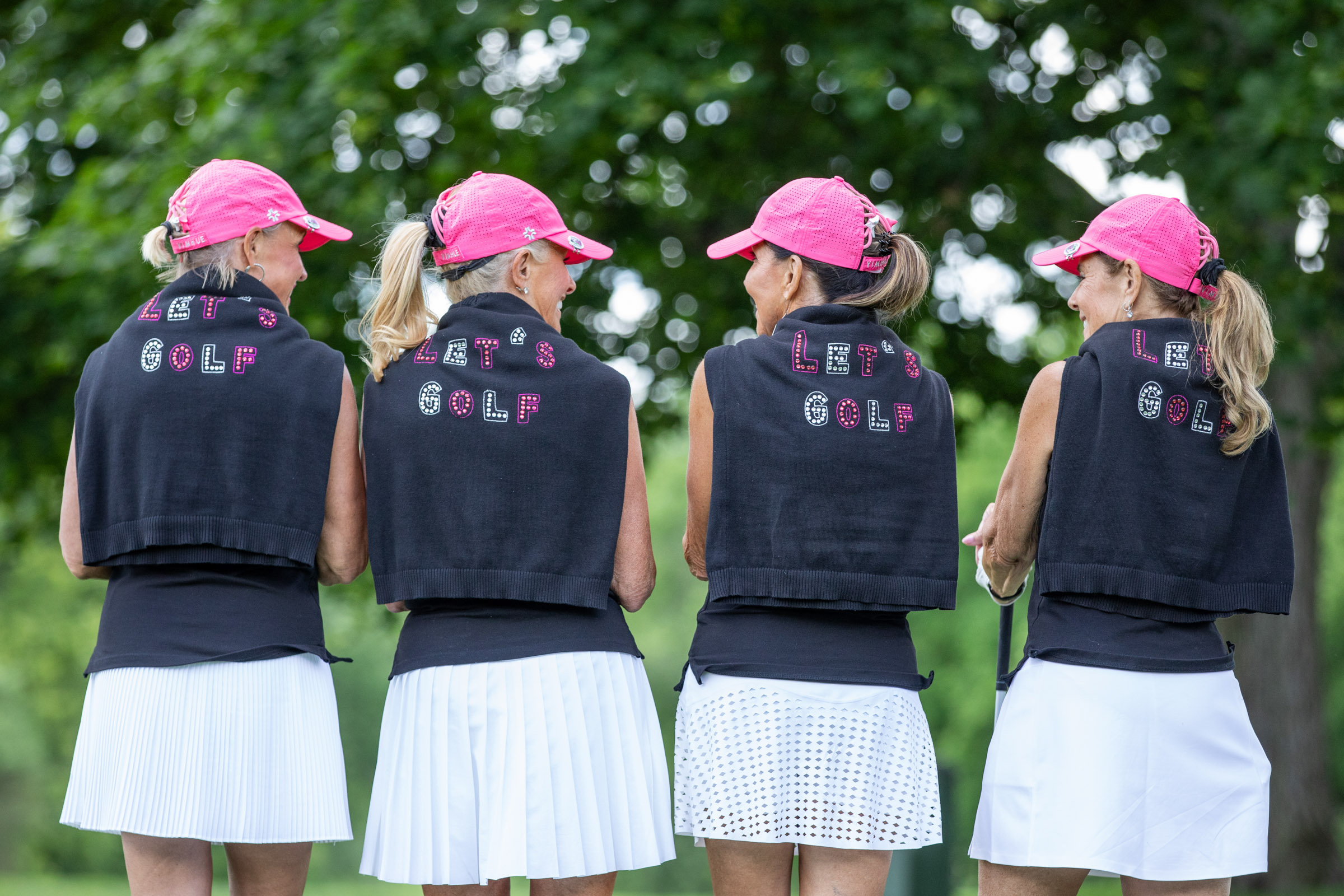 Four young girls in pink caps and navy vests stand outdoors with trees in the background.