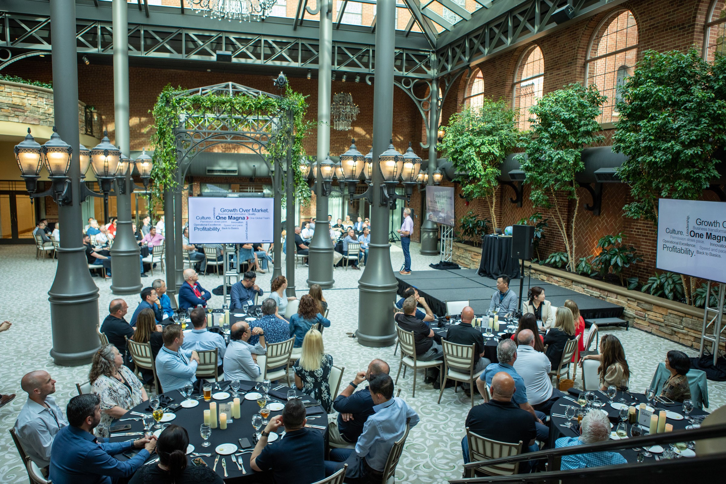 Indoor event with people seated at tables, a stage with a screen, and greenery in a spacious hall.