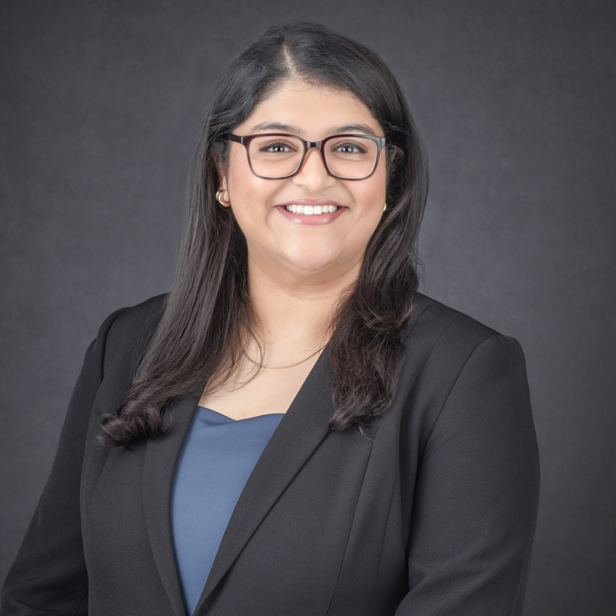 Smiling woman with glasses and long dark hair, wearing a black blazer and blue top, against a dark background.