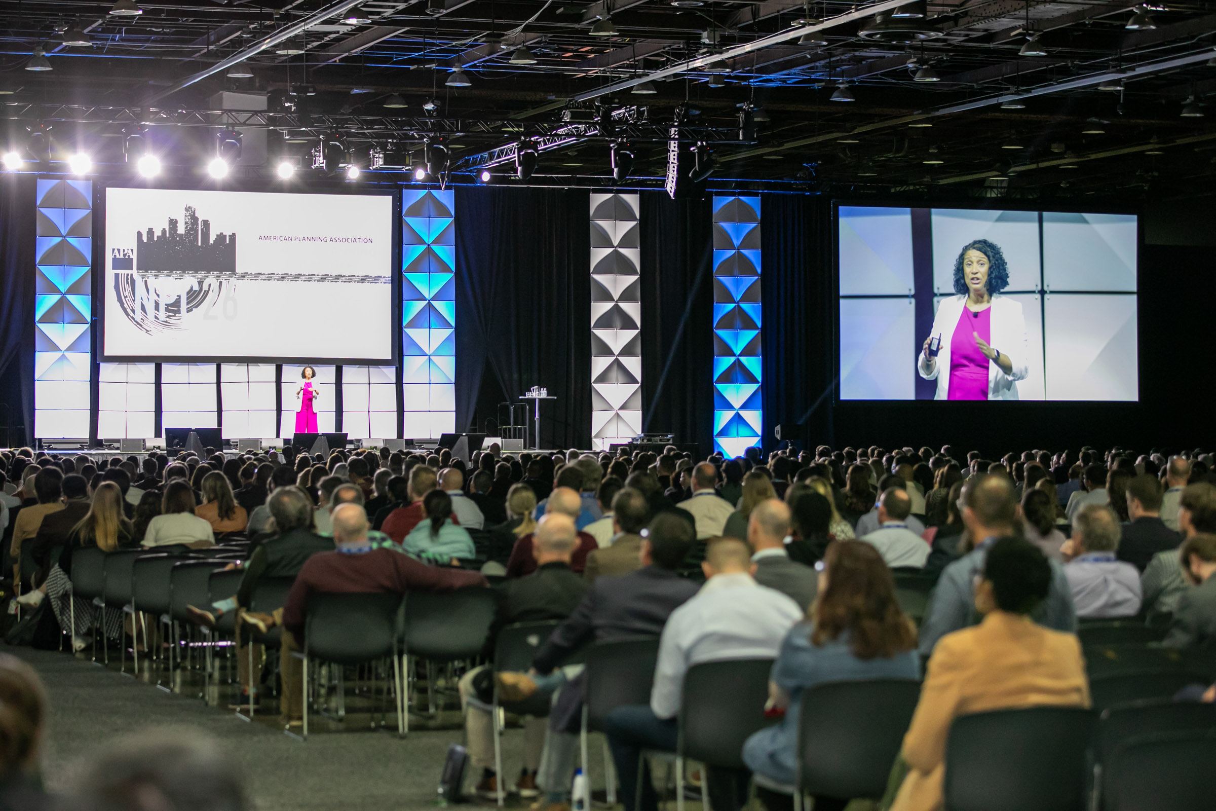 Large conference hall with a stage, large screens, and an audience watching a presentation.
