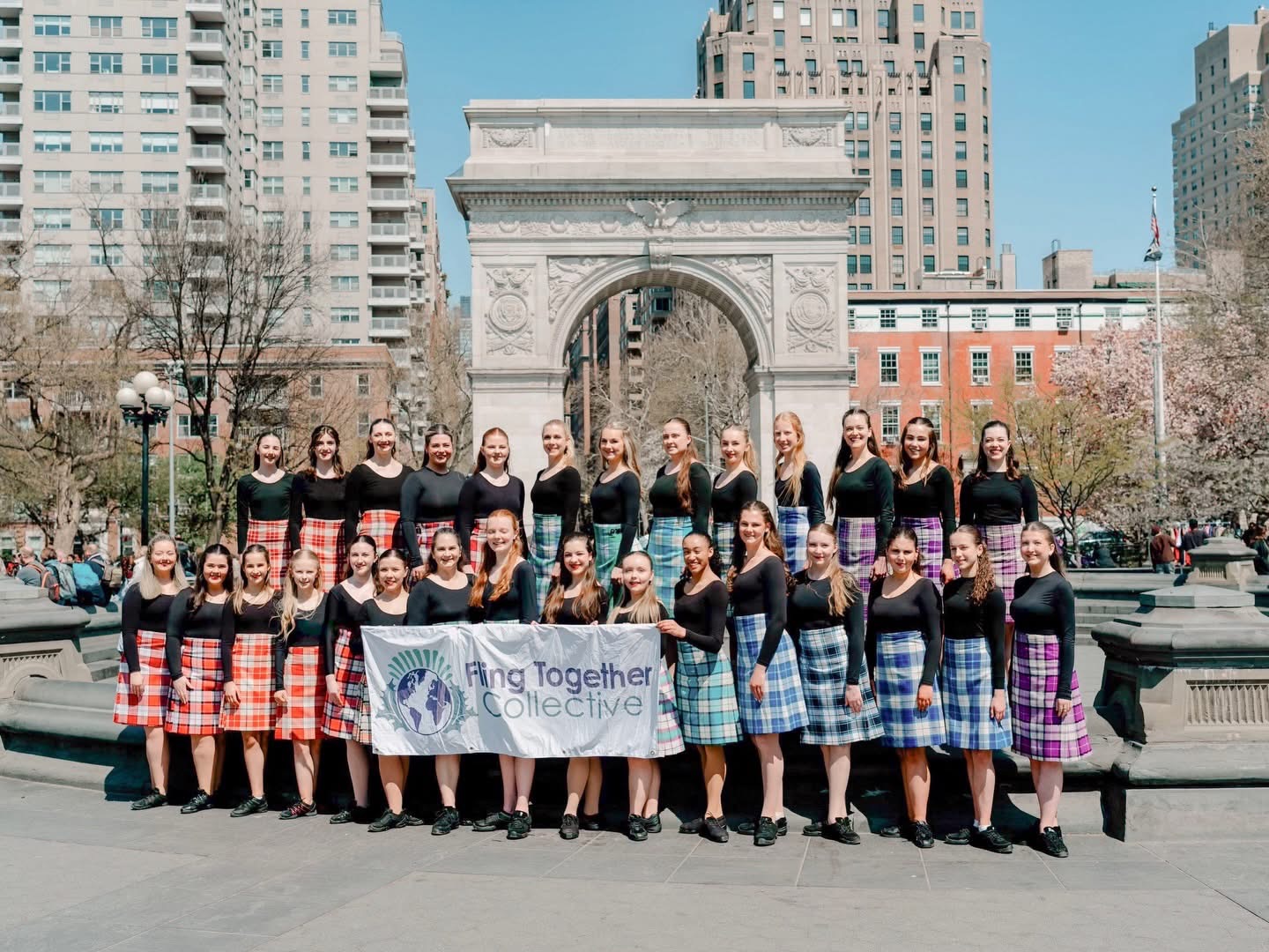 Group of women in plaid skirts holding a banner in front of a city arch, with tall buildings in the background.