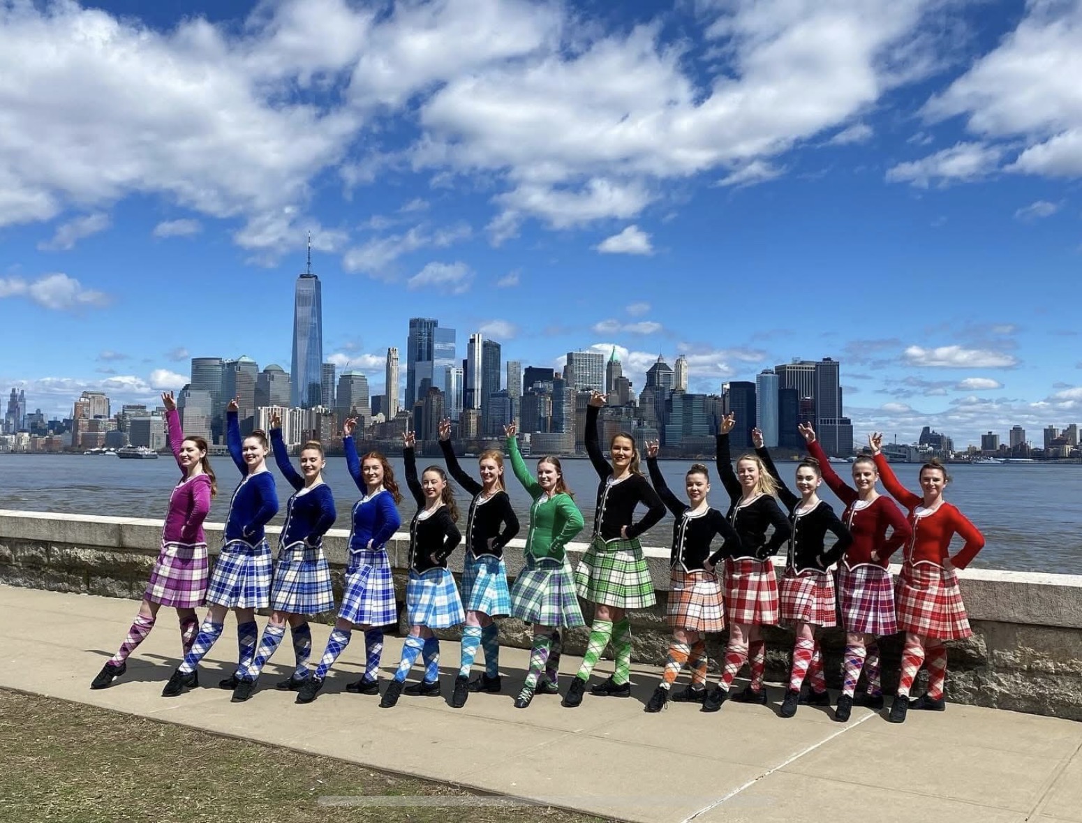 Group of people in colorful plaid skirts and black tops standing outdoors with city skyline and water in background.