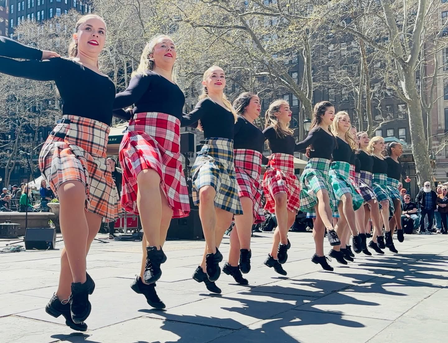 Group of women performing a synchronized dance outdoors, wearing black tops and plaid skirts, with trees and people in the background.