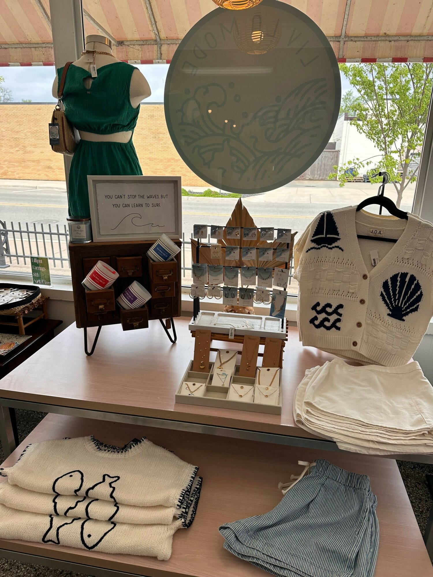 Display table with bags, clothing, and a sign, near a window with trees outside, in a retail setting.