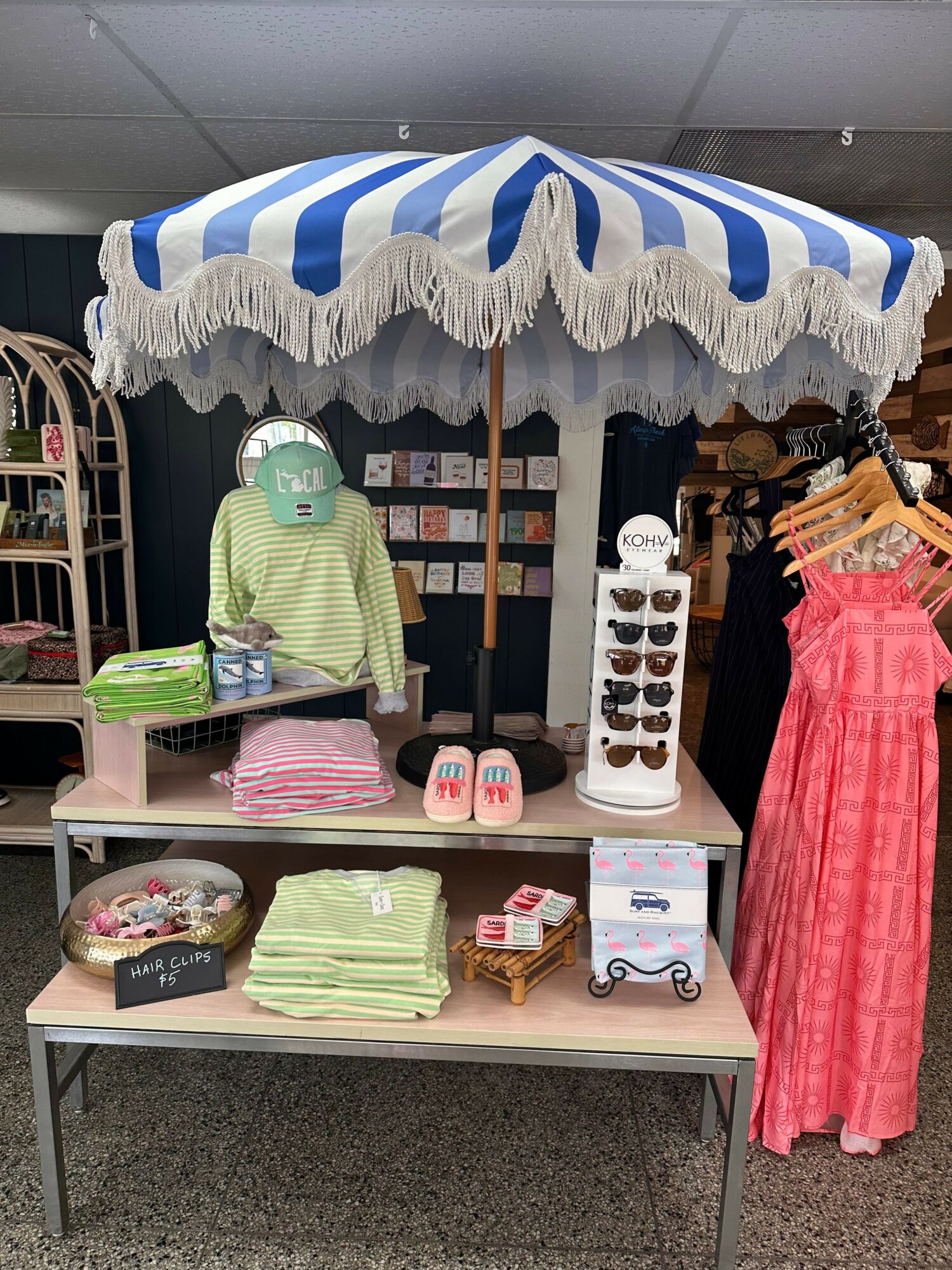 Display table with striped umbrella, clothing, and accessories, arranged on shelves and a small table, in a retail setting.
