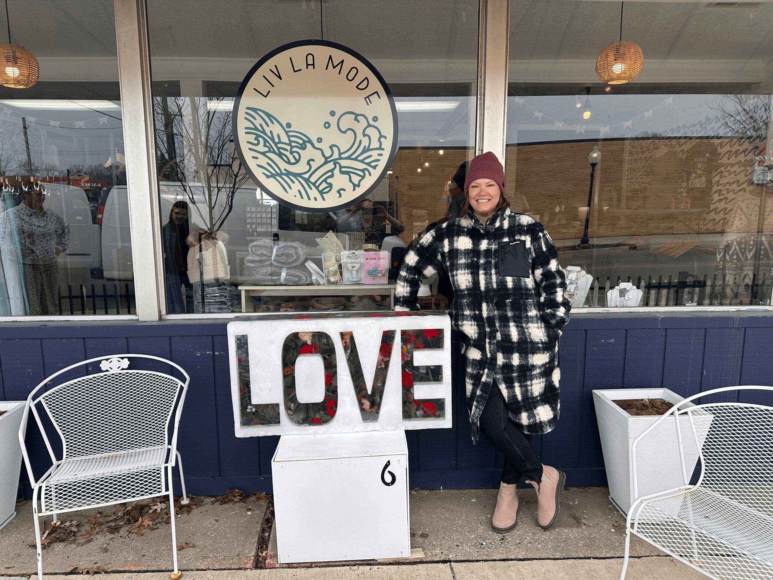 Person in black and white checkered coat and maroon hat standing outside a cafe with a LOVE sign, two white chairs, and a circular logo reading 'LIV LA MODE'.