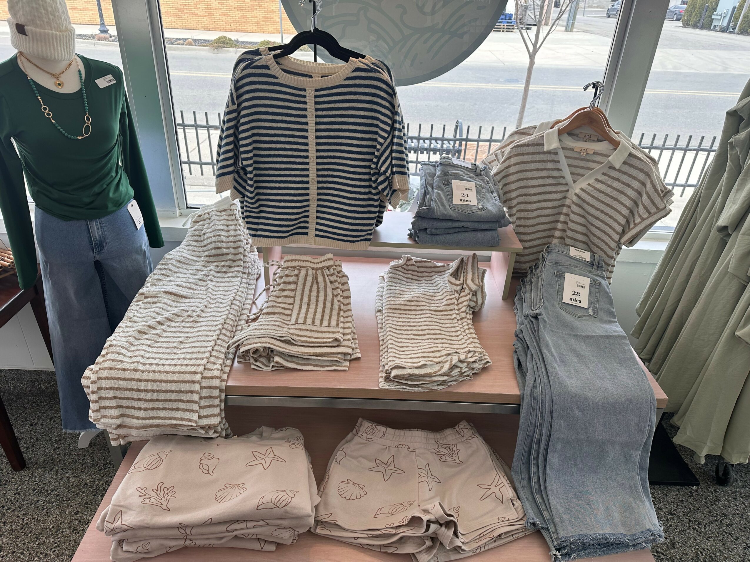 Display of striped and solid-colored clothing, including shirts, pants, and folded garments on a table near a window.