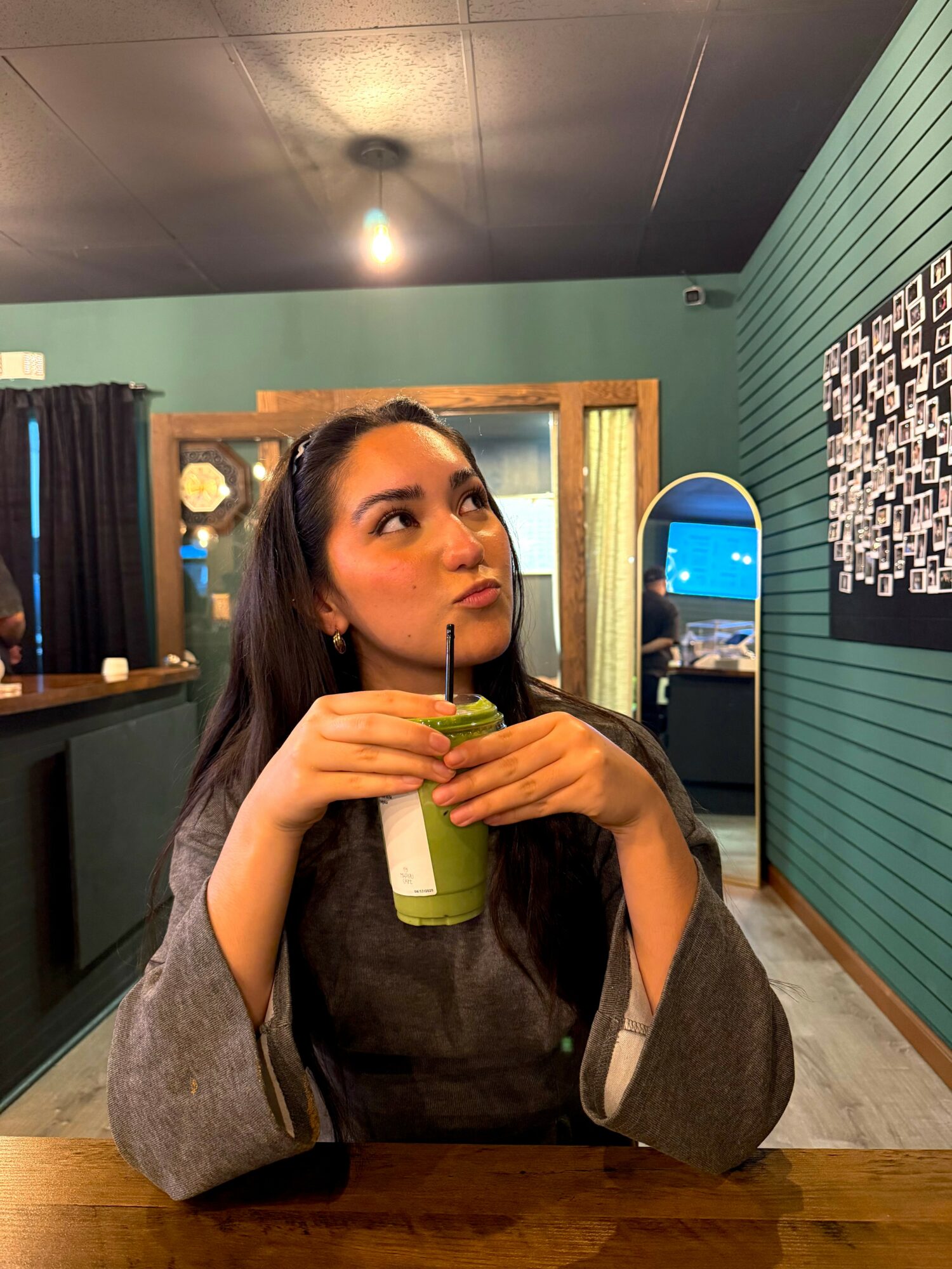 Woman sitting at a table holding a green drink with a straw, indoors with teal walls and a window behind her.