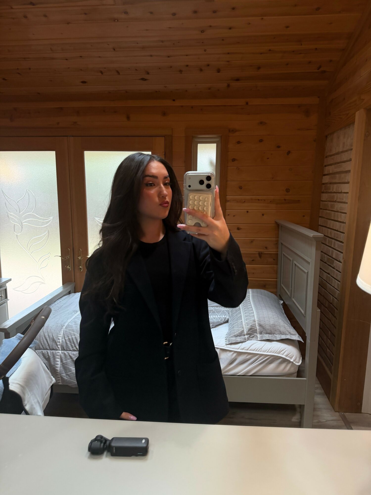 Woman taking a mirror selfie in a wooden-paneled room with a bed and window behind her.