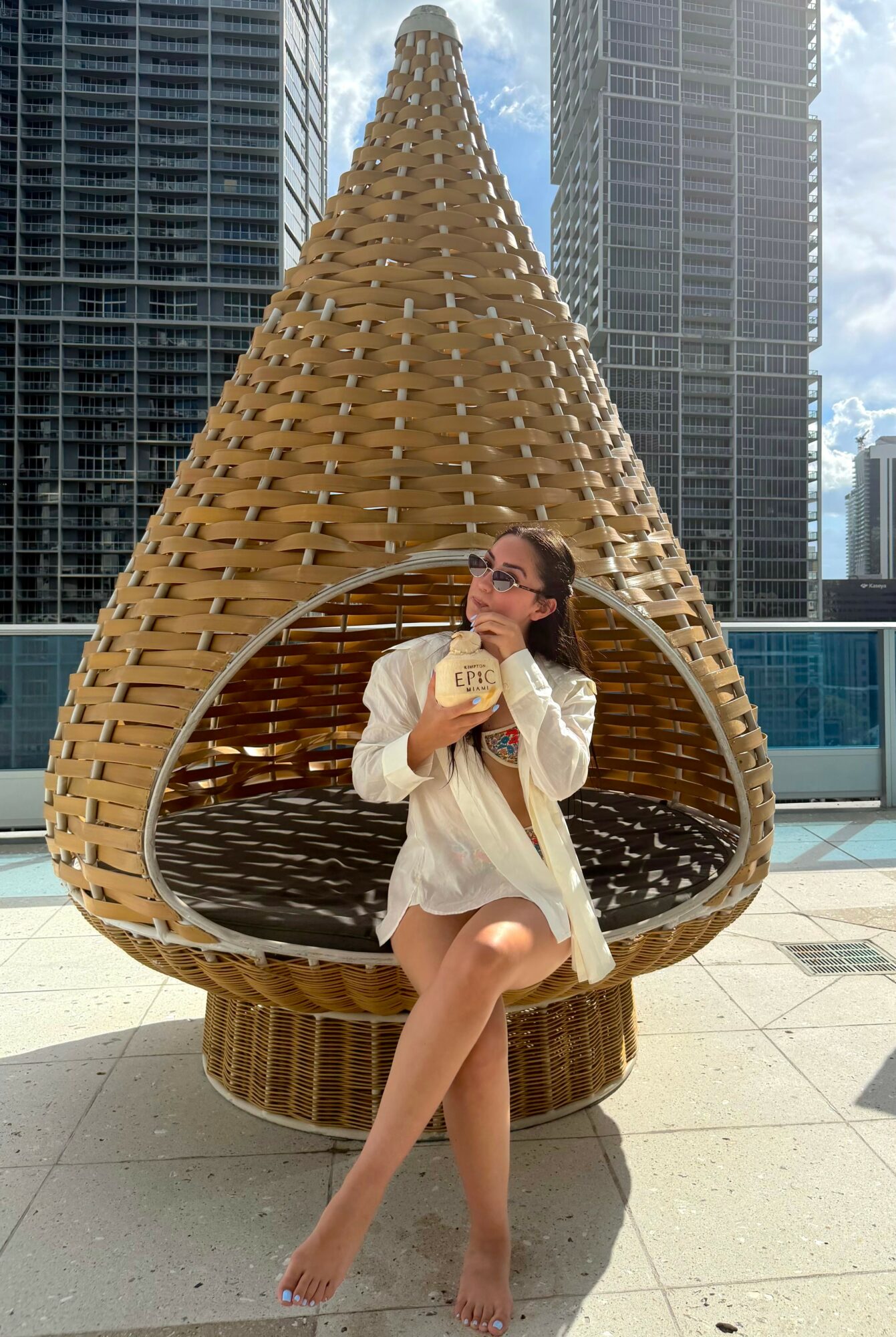 Woman sitting inside a teardrop-shaped wicker structure on a rooftop with tall buildings in the background.