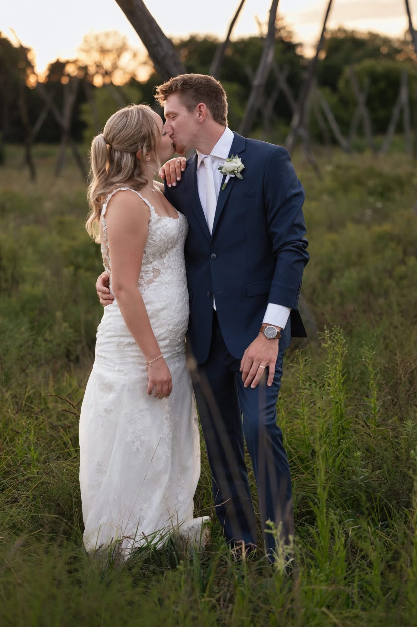 A bride and groom stand close together outdoors, about to kiss, with trees and grass in the background.