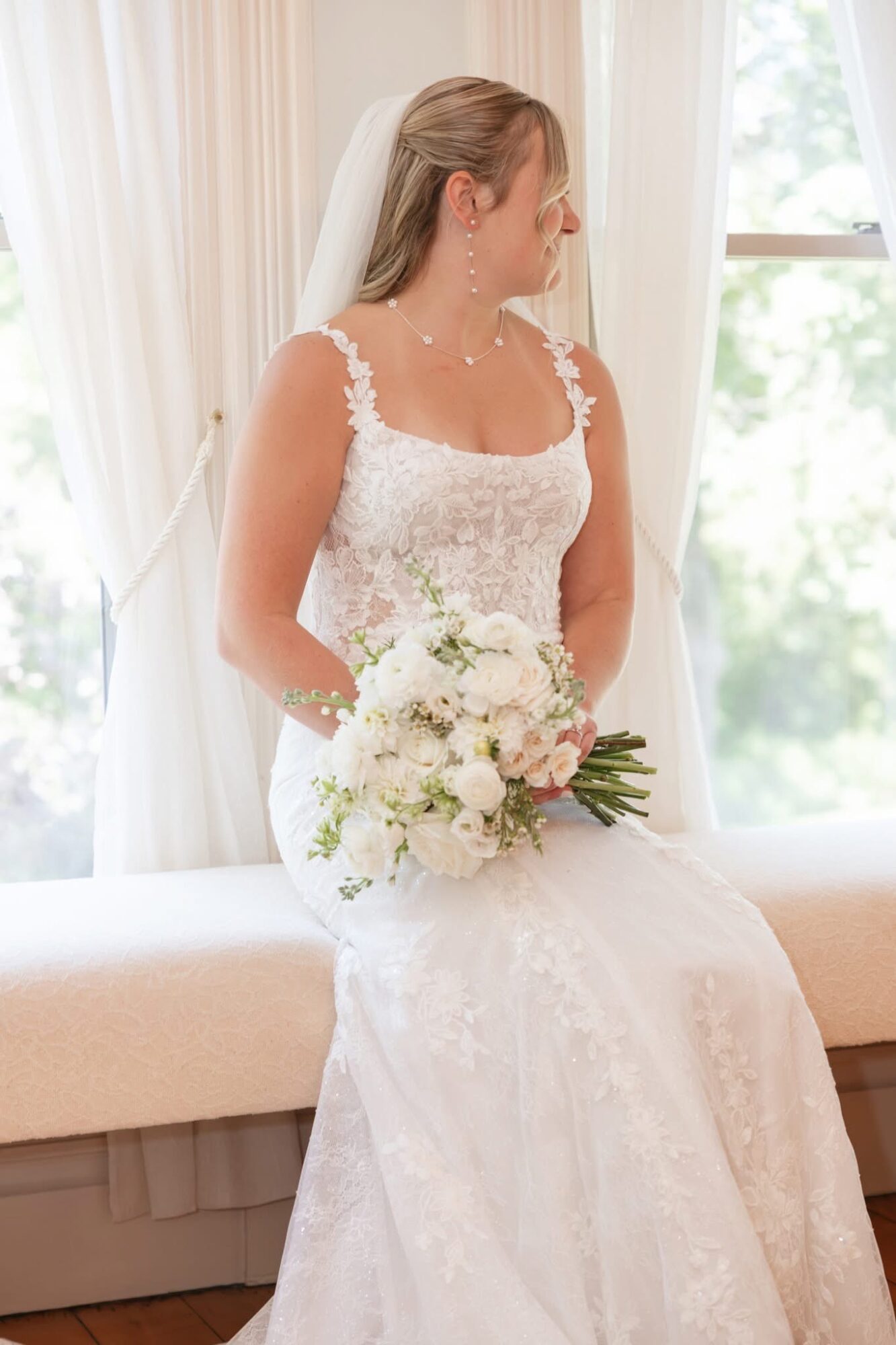 Bride in wedding dress holding a bouquet, sitting indoors near a window with curtains, looking to the side.