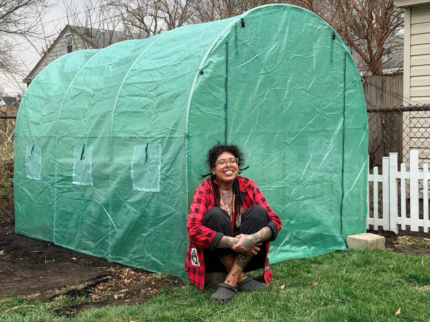 Person sitting outside a green greenhouse, smiling, with a white picket fence and house in the background.
