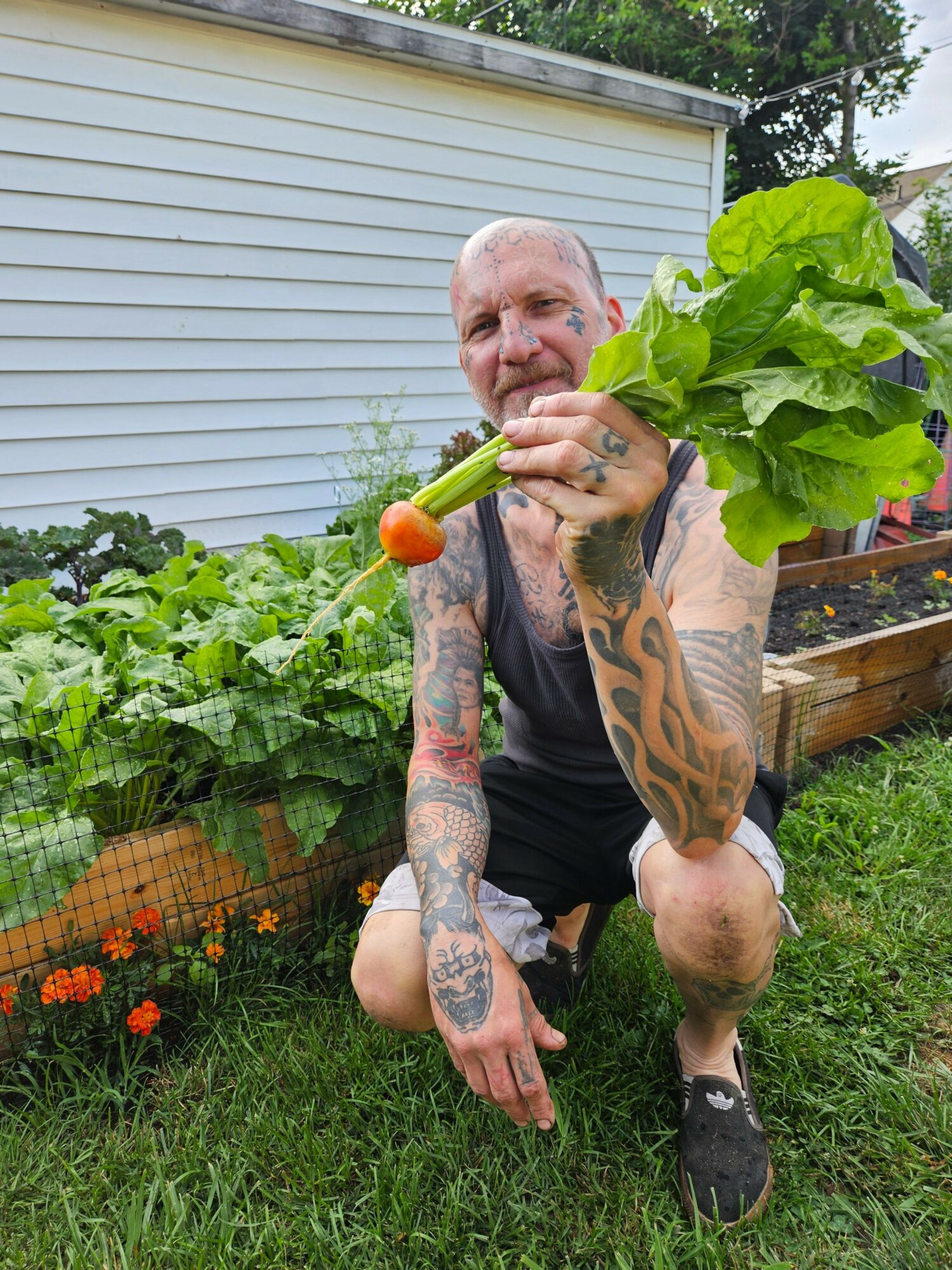 Person with tattoos holding a bunch of leafy greens and a carrot in a garden with plants and flowers.