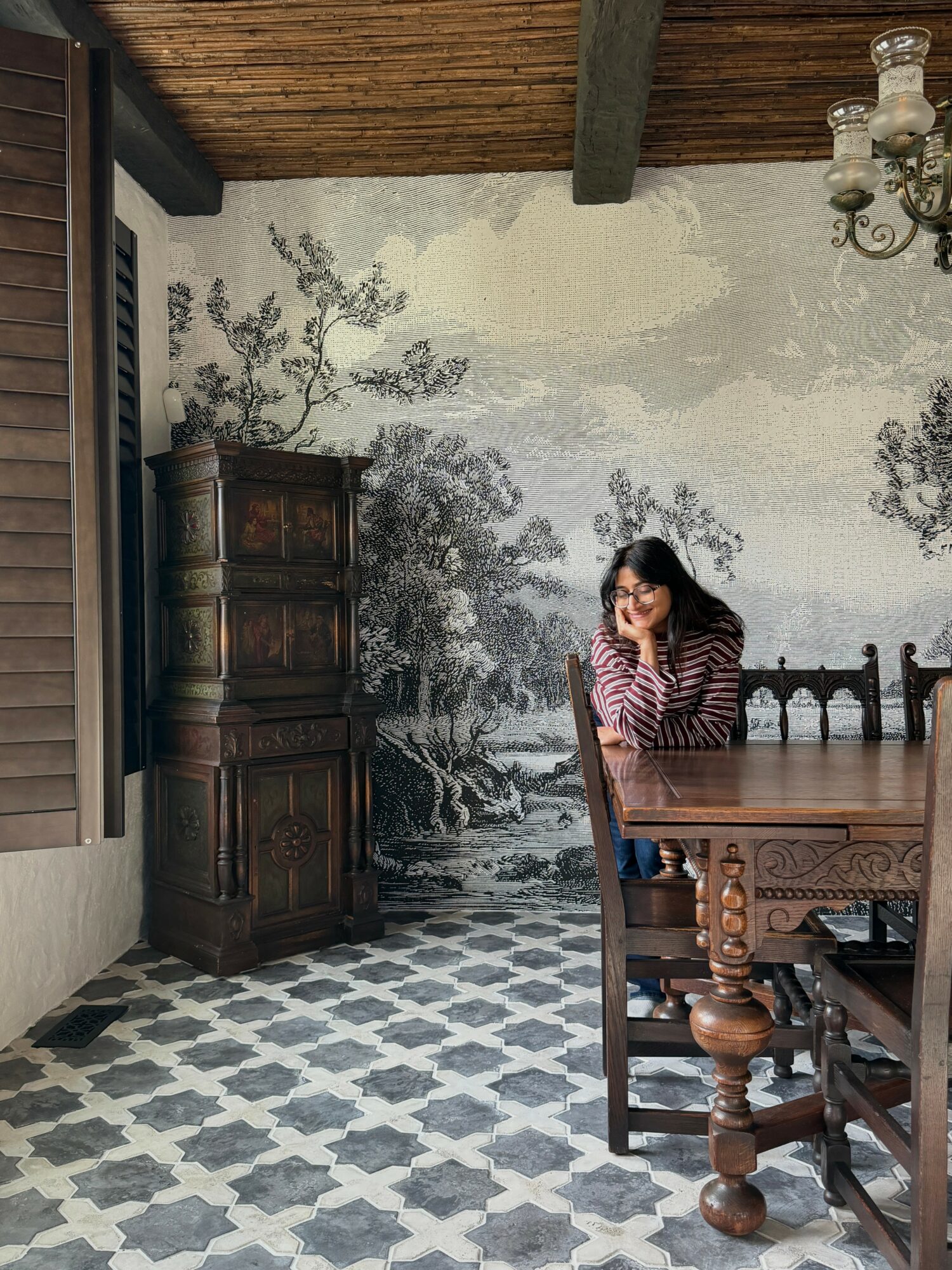 Woman sitting at a wooden dining table in a room with a mural of trees on the wall, wooden ceiling, and patterned floor.