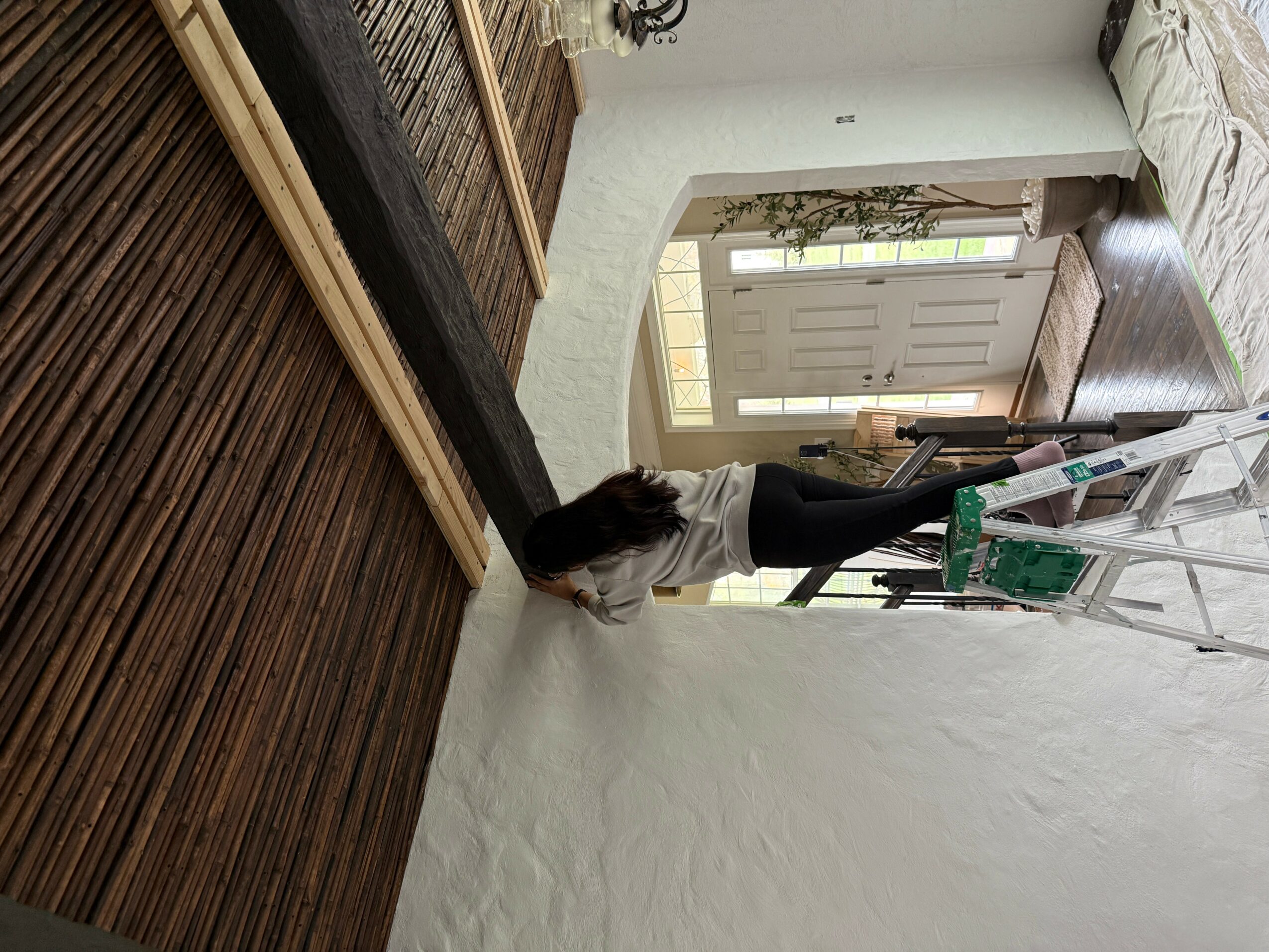 Person leaning over a white wall, with a ladder and a green object nearby, in a room with a door and window.