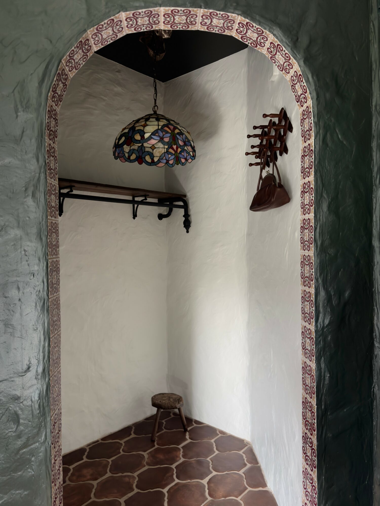 Small corner with hanging lamp, shelf, and coat hooks, with a stool on hexagonal tile floor, arched doorway with decorative border.