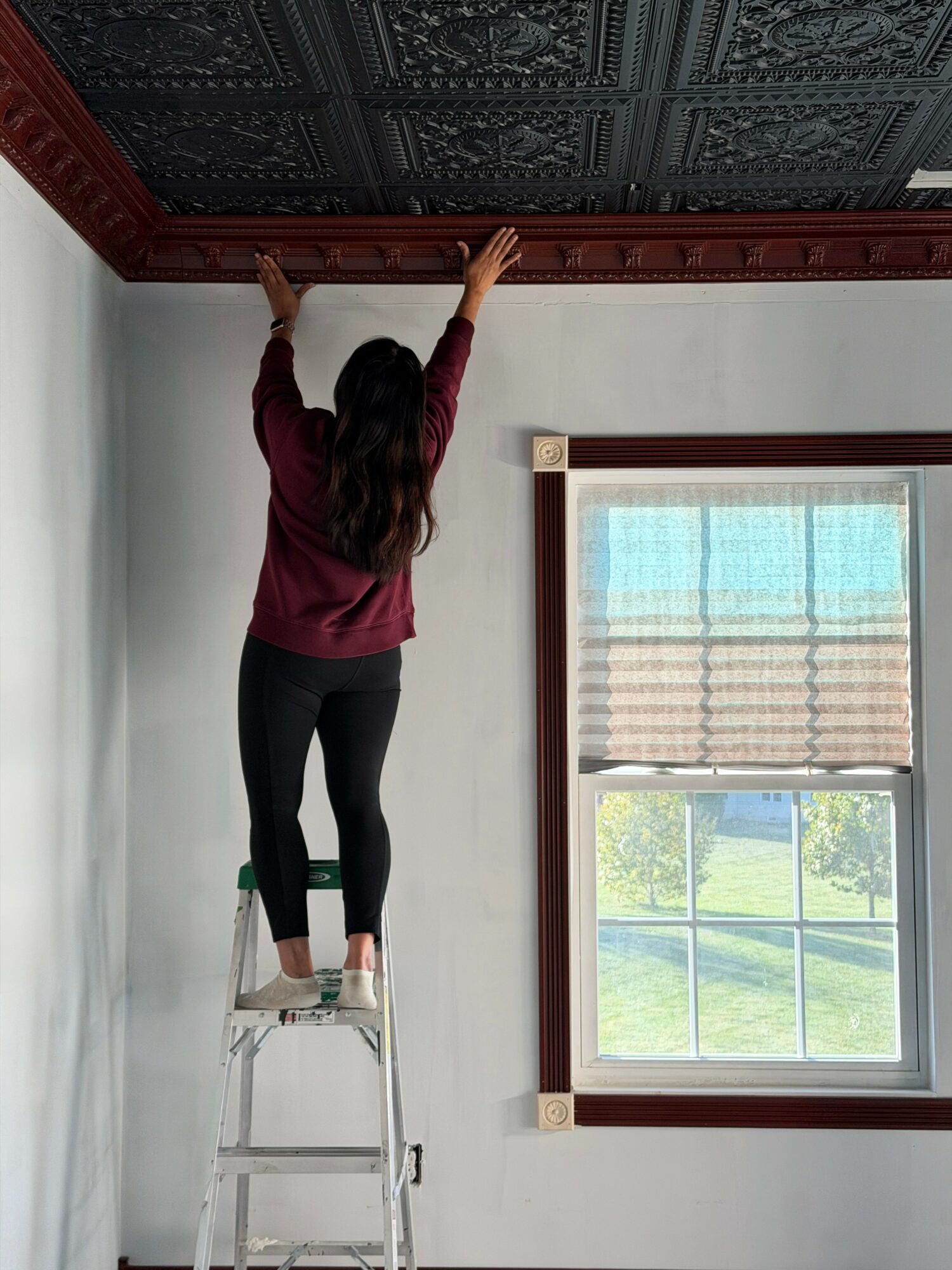 Person standing on a ladder reaching up to the ceiling, with a window and blinds nearby.