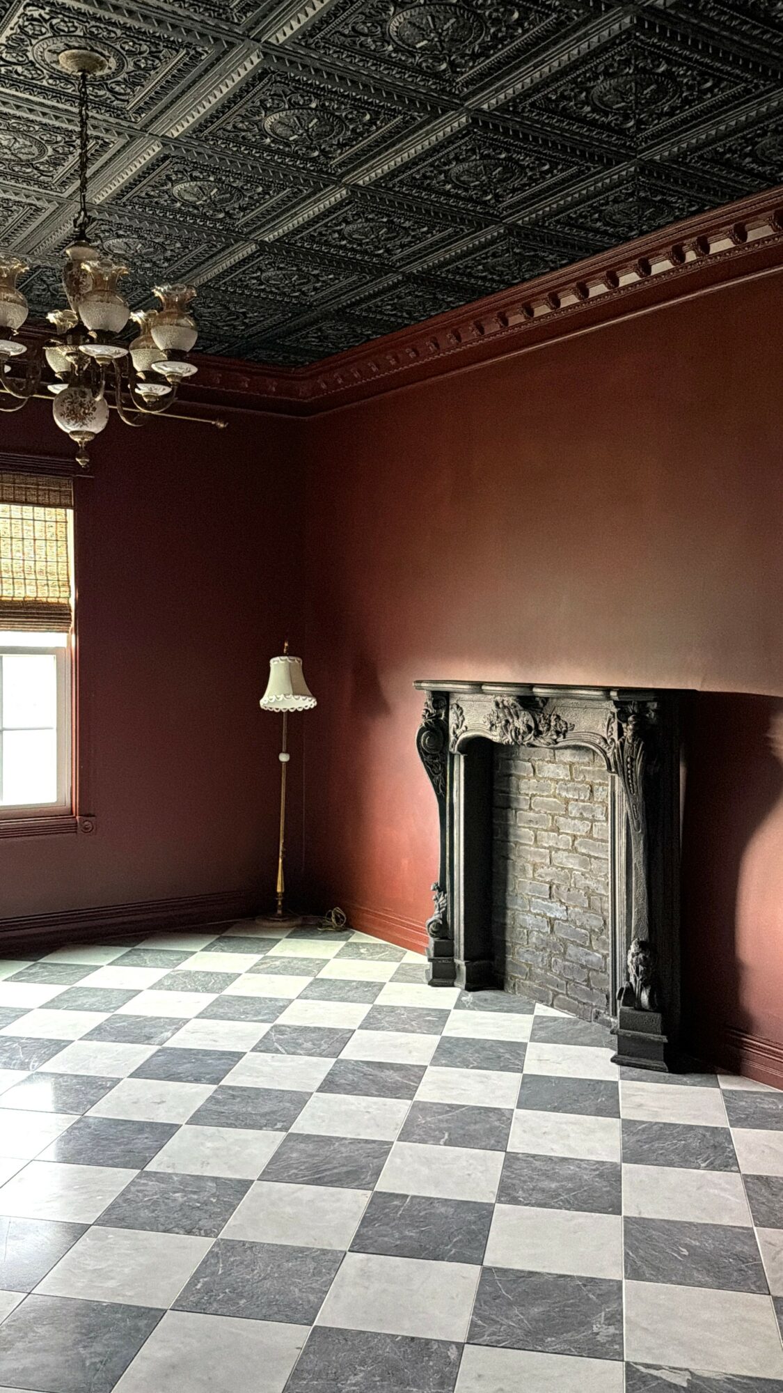 Room with checkered floor, ornate fireplace, red walls, chandelier, and window with blinds.