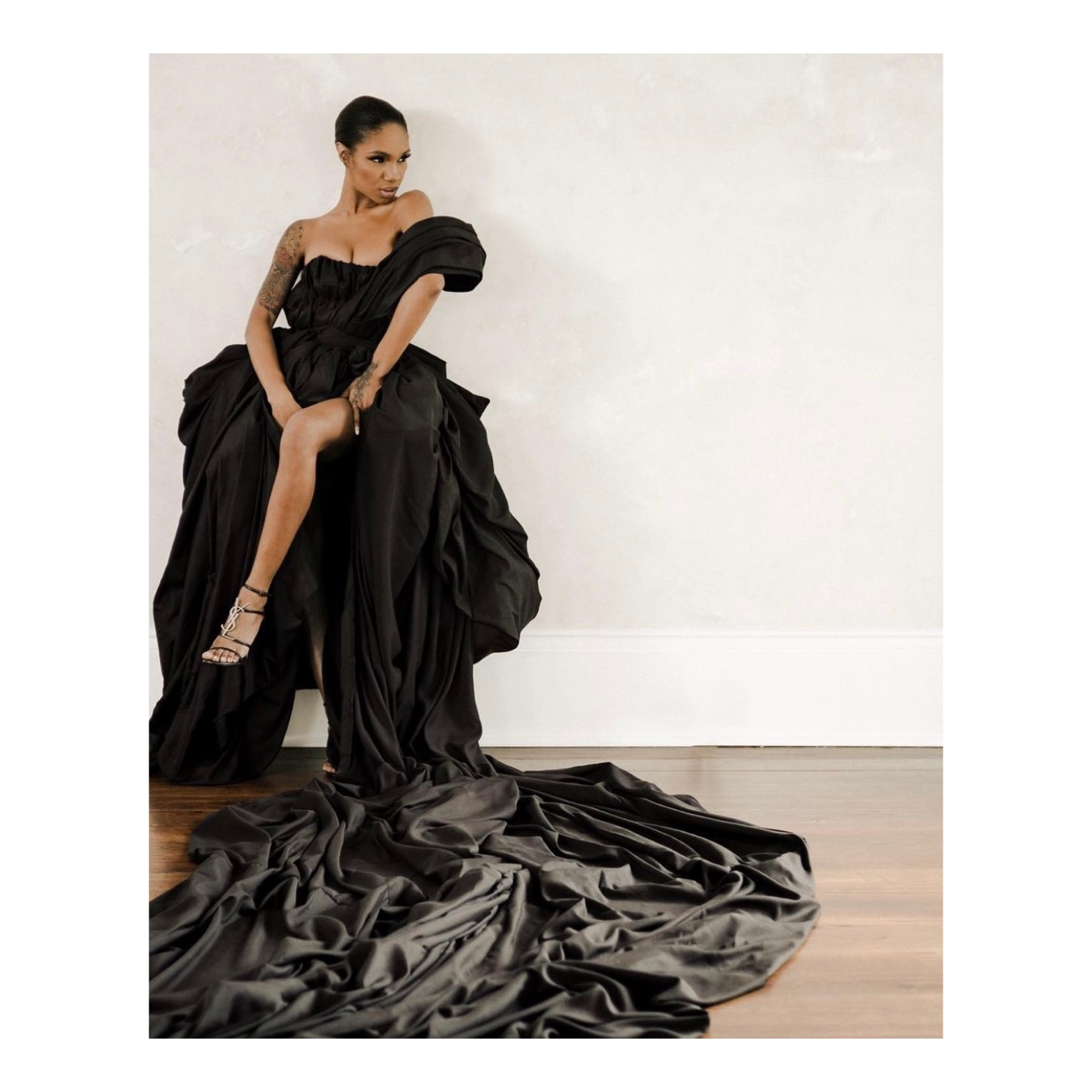 Woman in a black gown with a long train, sitting on a chair, against a plain white wall.