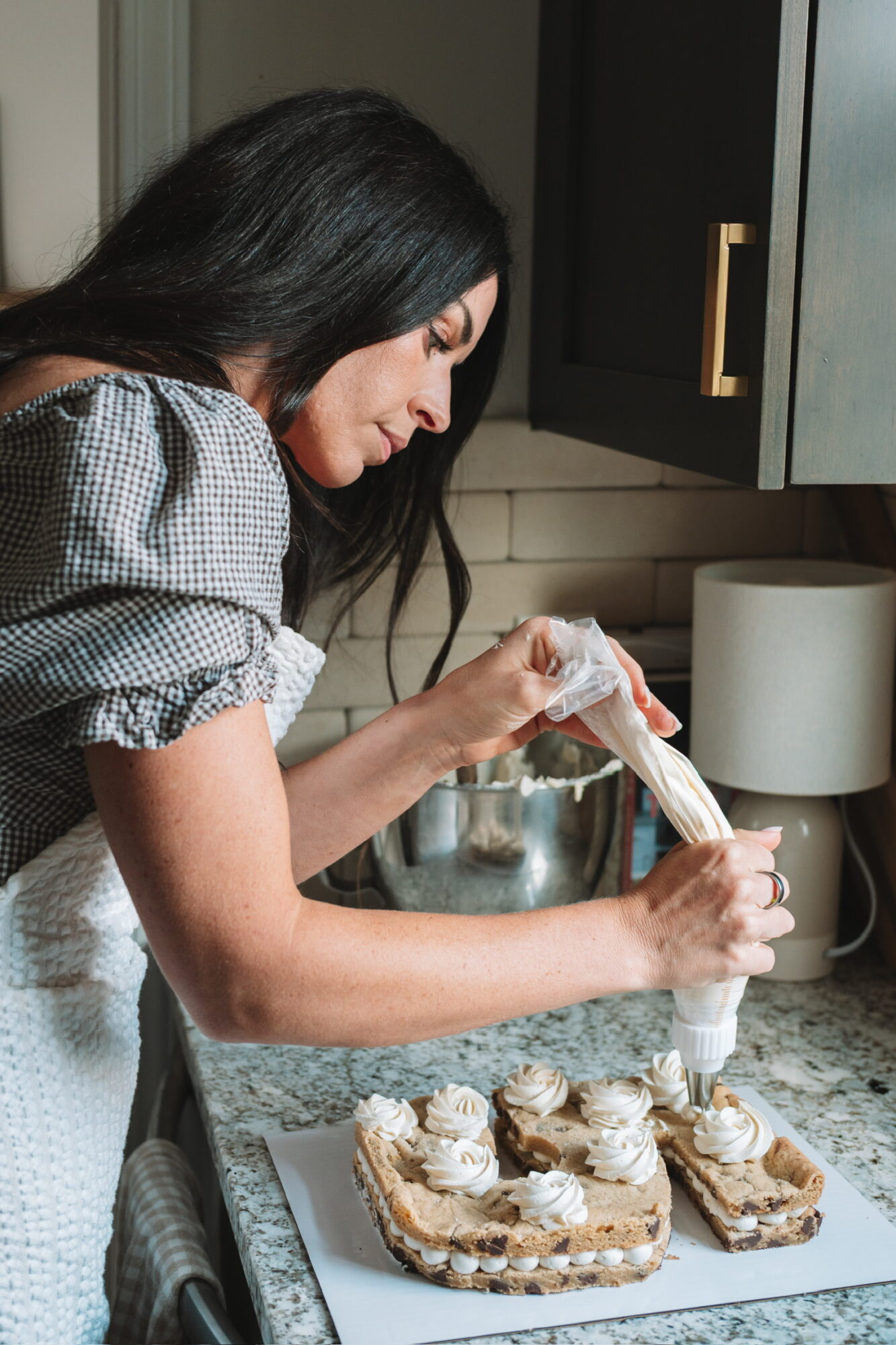 Woman piping frosting onto cupcakes on a kitchen counter, with a cabinet and lamp in the background.