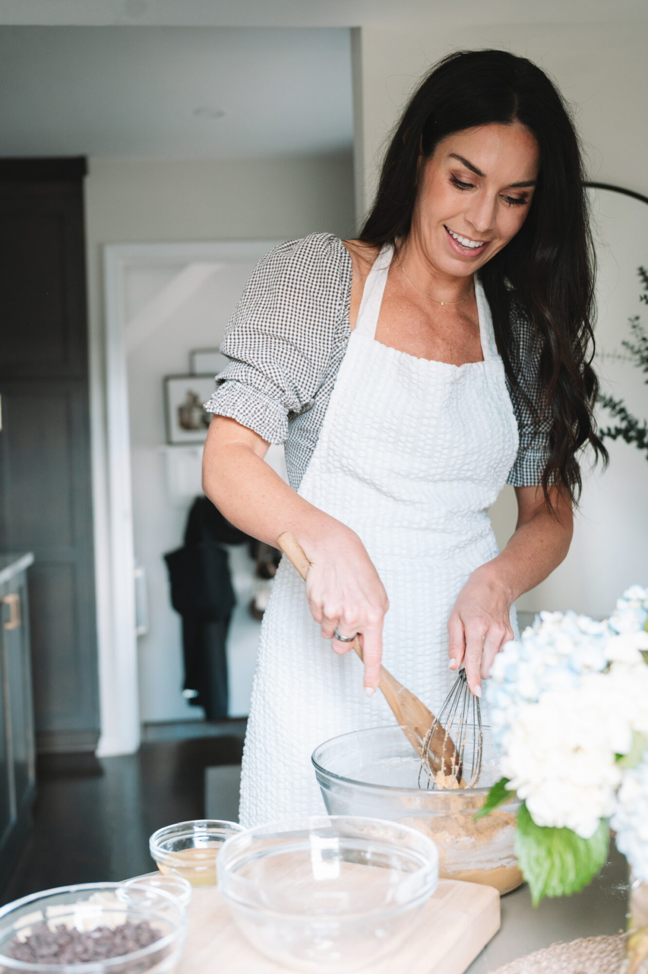 Woman mixing ingredients in a bowl, wearing an apron, in a kitchen with flowers on the counter.