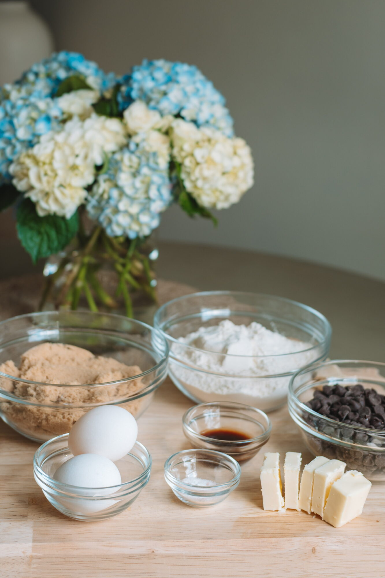 Vase with blue and white hydrangeas, bowls of eggs, flour, chocolate chips, vanilla, and butter on a wooden table.