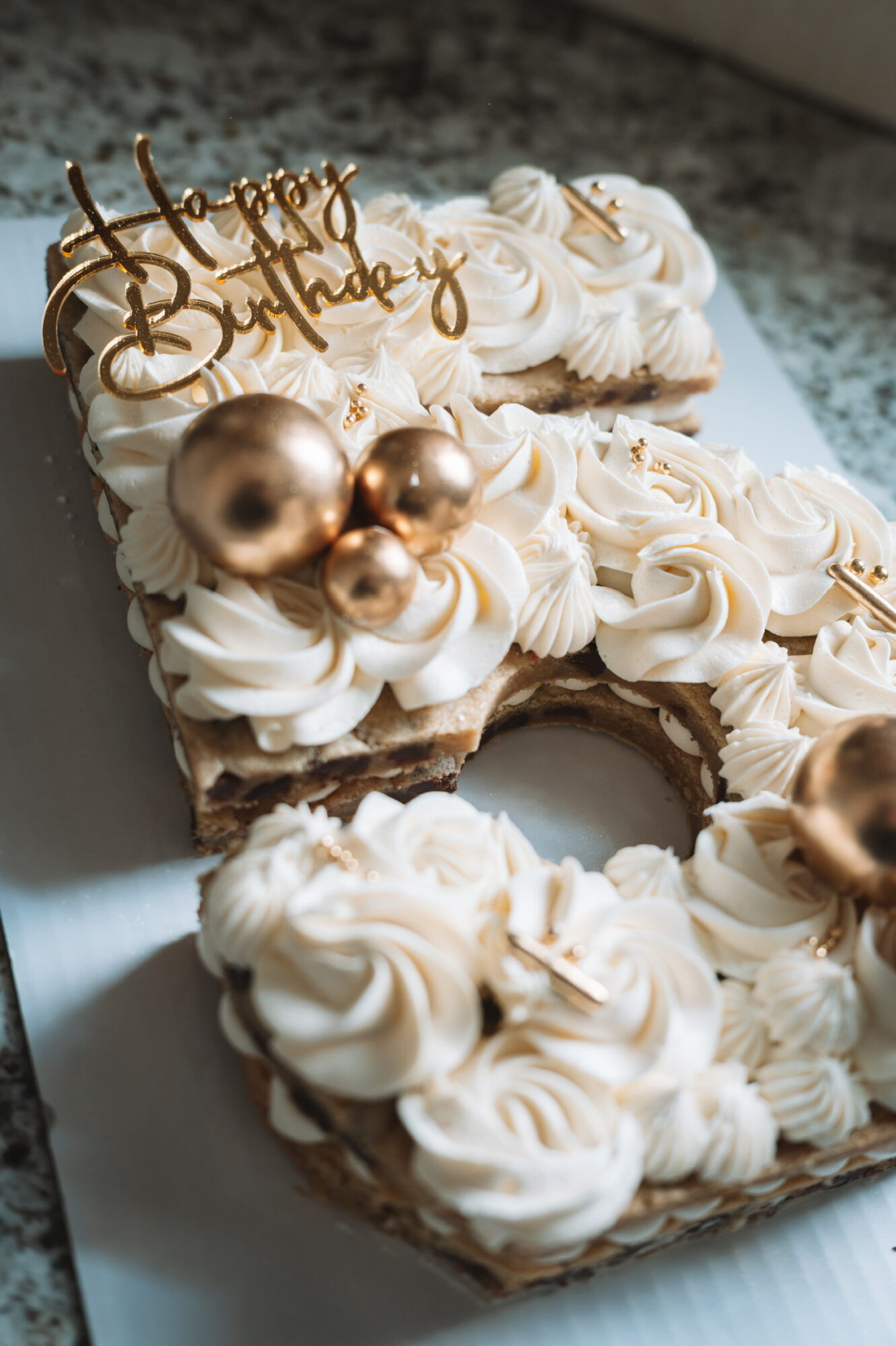 Celebration cake decorated with white frosting, gold ornaments, and a 'Happy Birthday' topper, on a white surface.