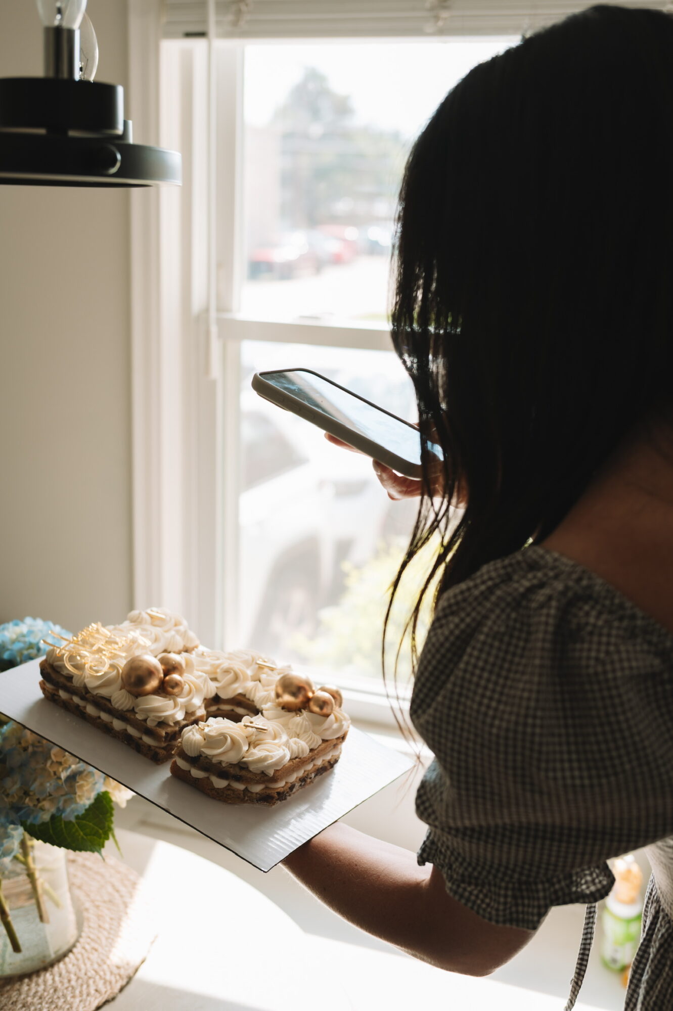 Person holding a plate with a cake, looking at a smartphone, in a bright room with a window